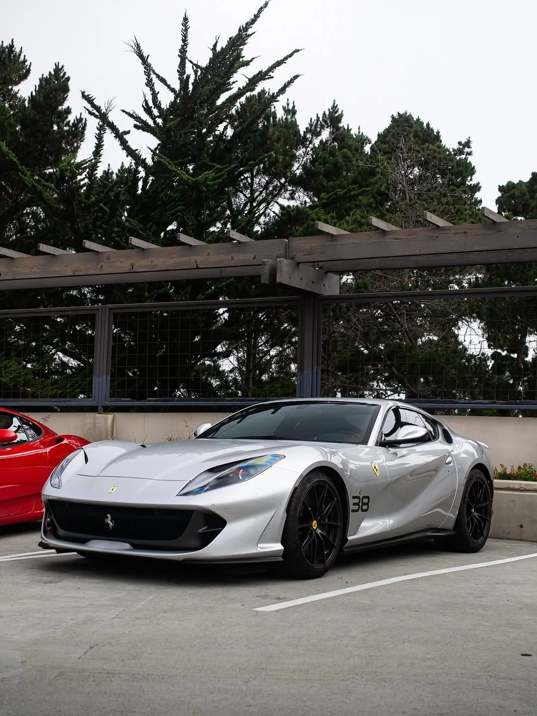 A silver Ferrari sports car parked in a lot next to a red car, with a background of trees and a wire fence.