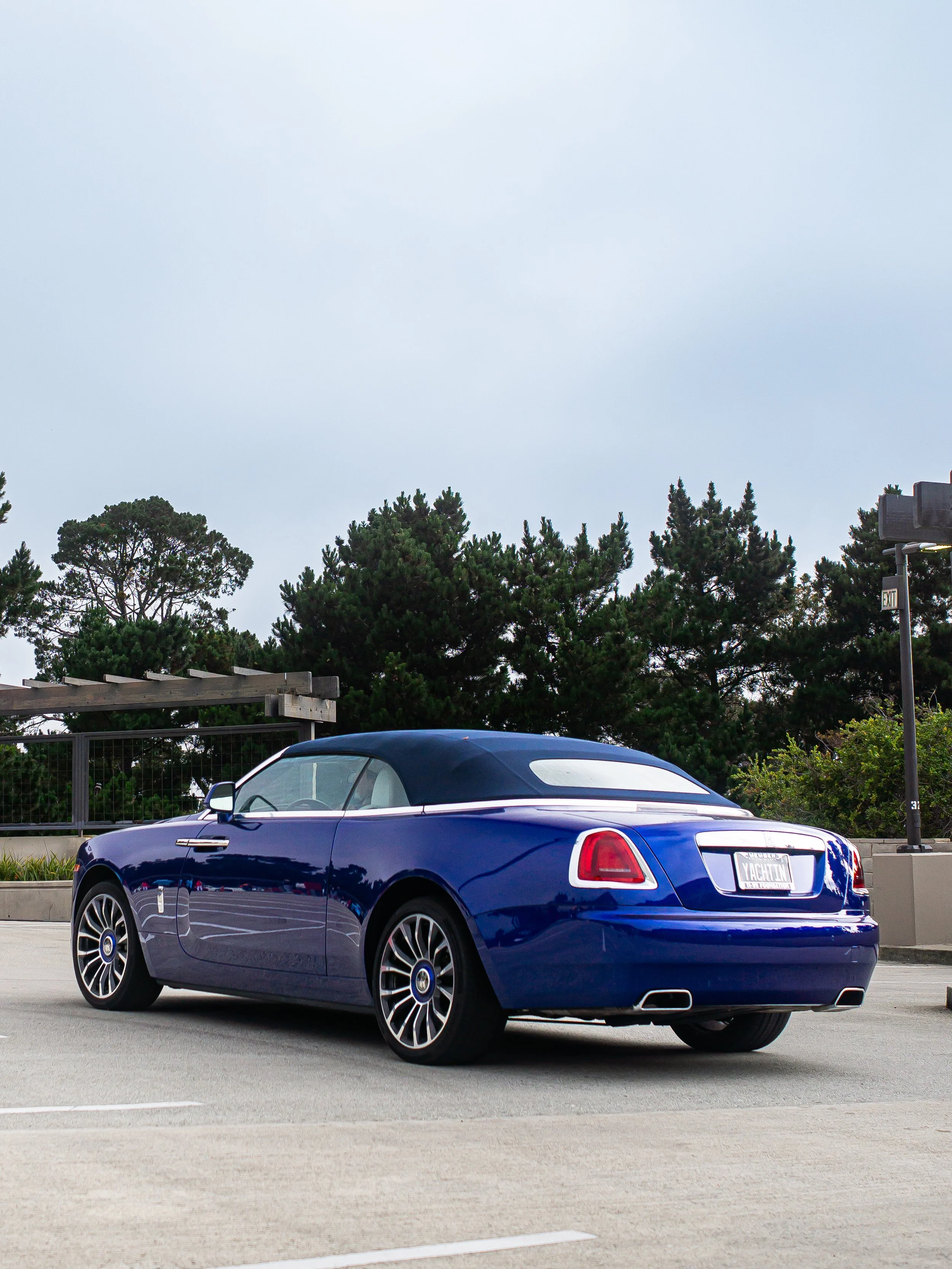 Blue luxury convertible car parked in an outdoor parking lot with trees in the background.