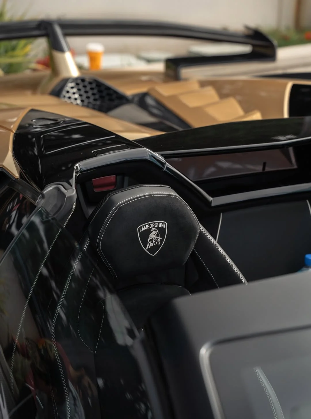 Close-up of a black Lamborghini steering wheel with white stitching, showing the Lamborghini logo, inside a luxury sports car.