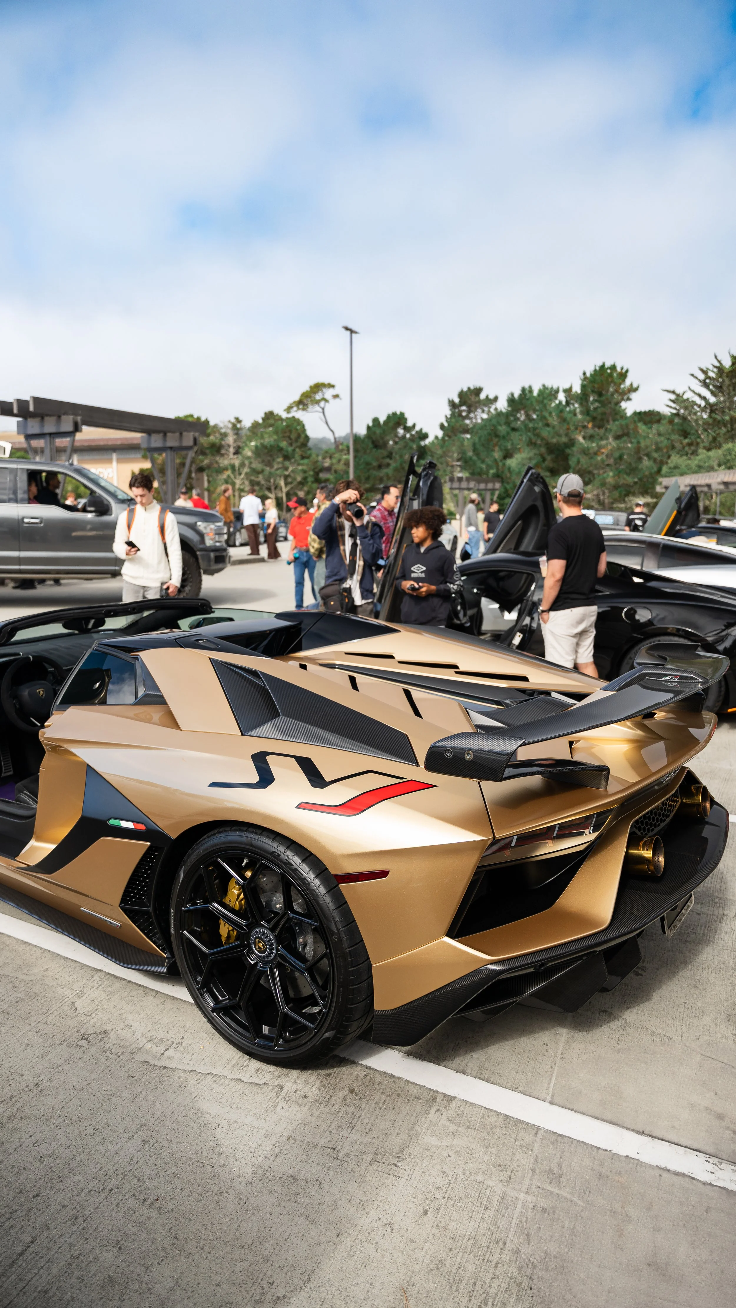A gold Lamborghini Sian at a car show, surrounded by people, with trees and a clear sky in the background.
