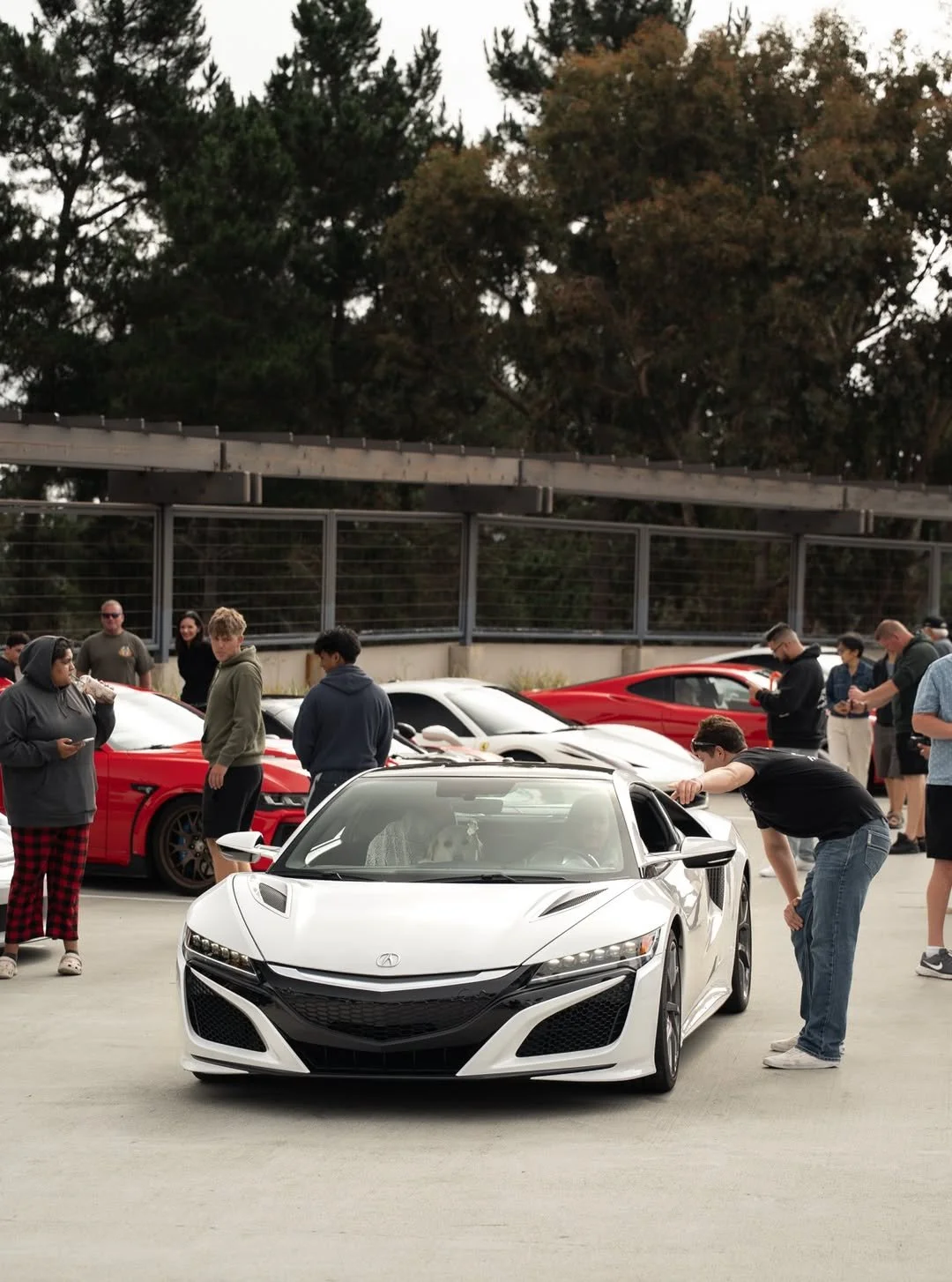 White Acura sports car at a car meet, surrounded by other cars and people admiring and taking pictures.