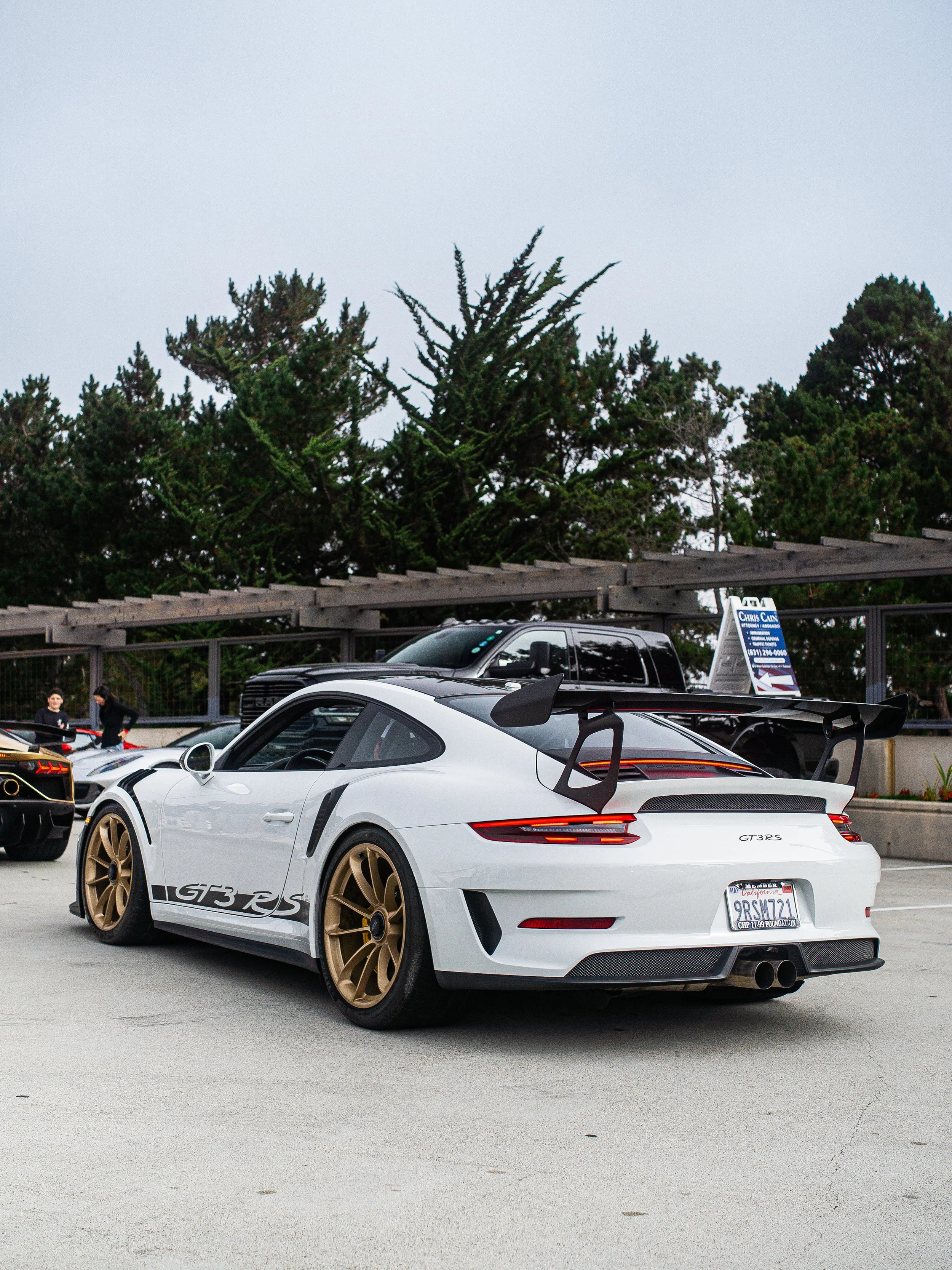 A white Porsche 911 GT3 RS with a large rear wing, gold wheels, and black accents is parked in an outdoor lot. There are other cars and people in the background.