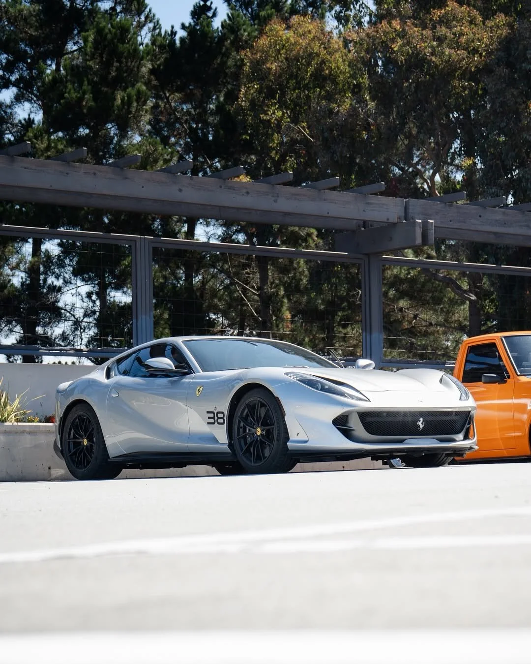 A silver Ferrari sports car with black wheels and a racing number 38 on the side, parked next to an orange vehicle on a concrete surface, with trees and a wooden pergola in the background.