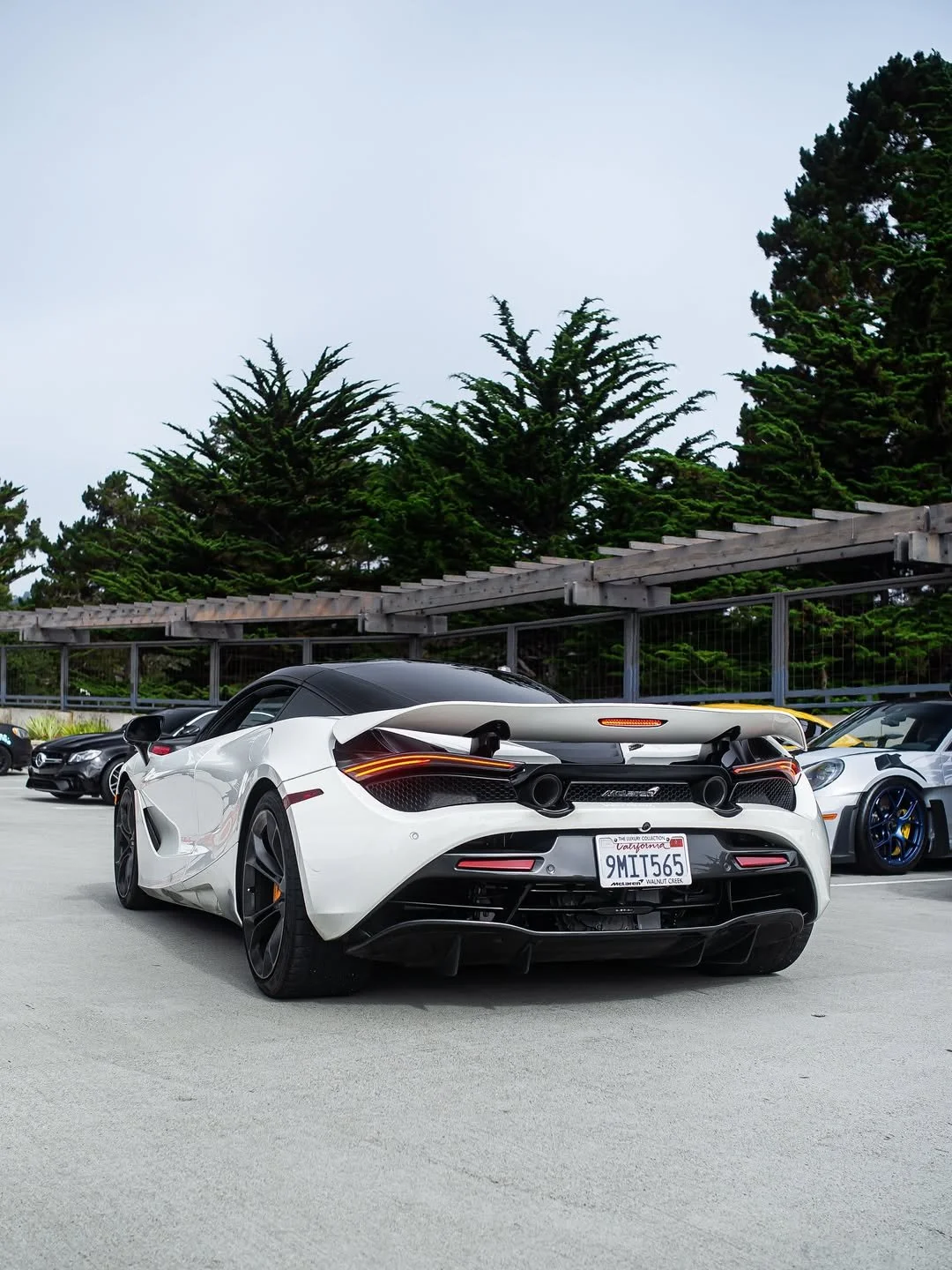 A white McLaren sports car with a large rear spoiler parked in a lot with other cars, trees, and a fence in the background.