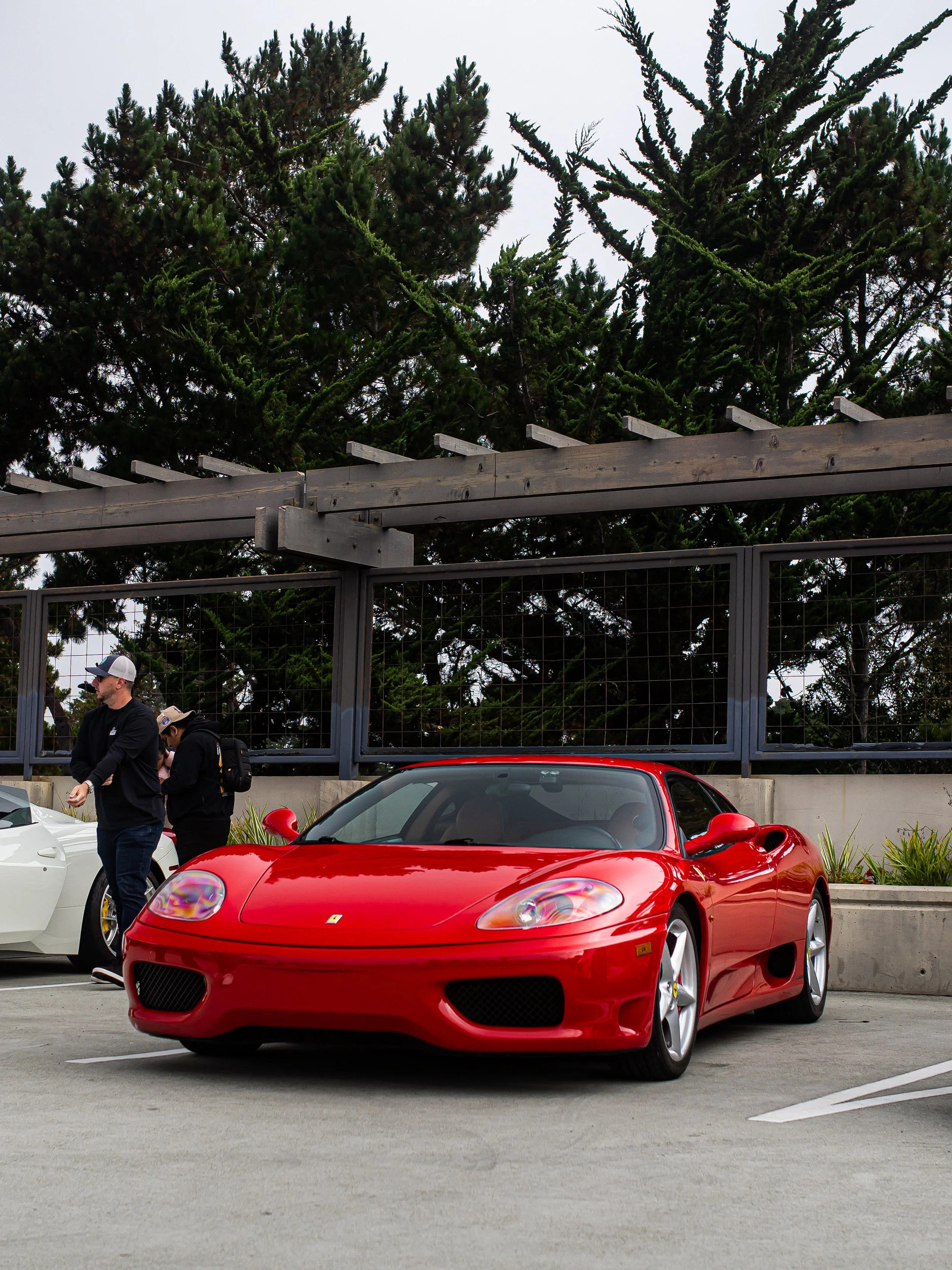 Red Ferrari sports car parked in a parking lot with two people standing nearby, behind a concrete barrier and metal fence with tall trees in the background.