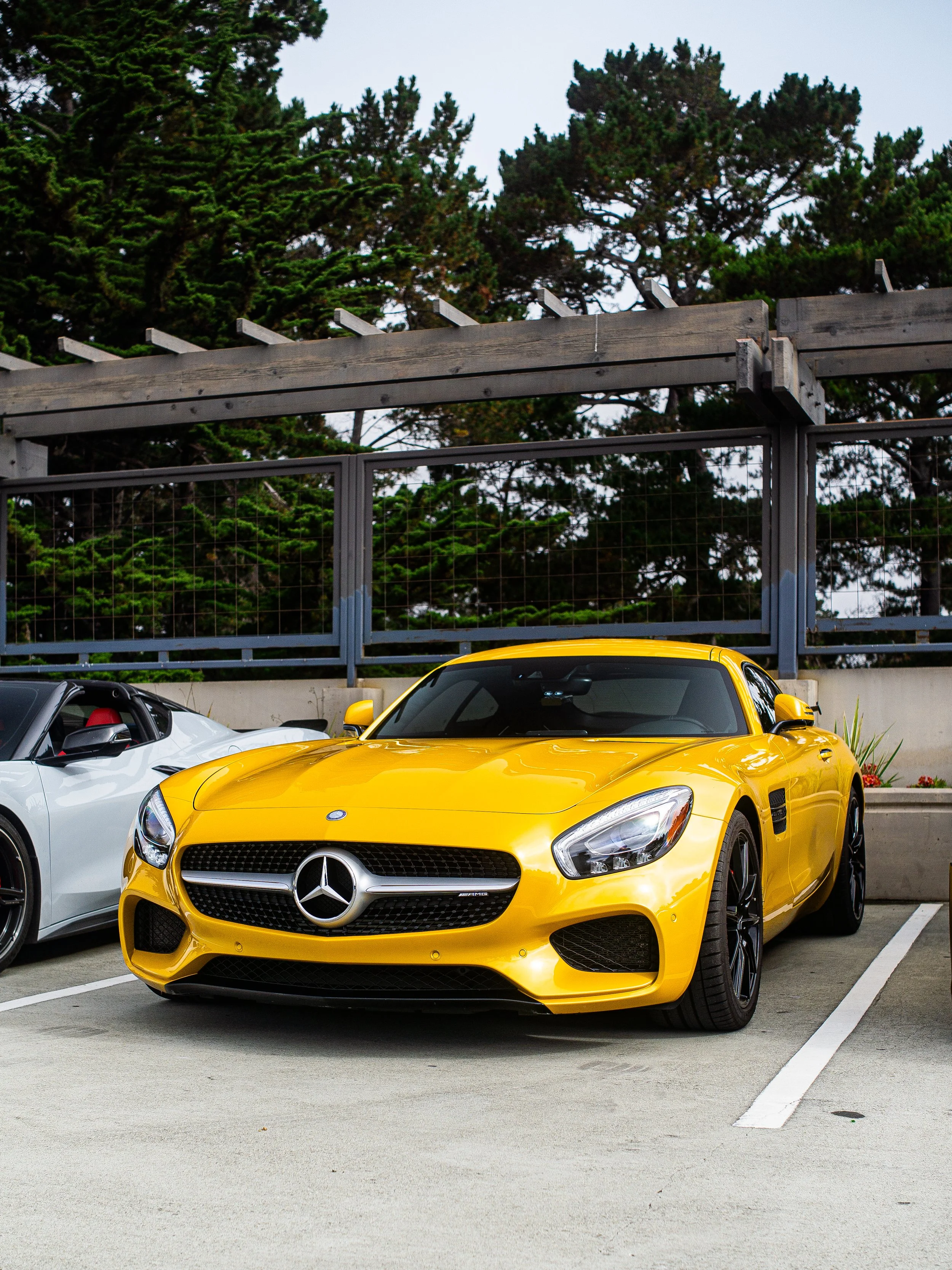 Yellow Mercedes-Benz sports car parked in a parking lot next to a white convertible, with trees and a fence in the background.