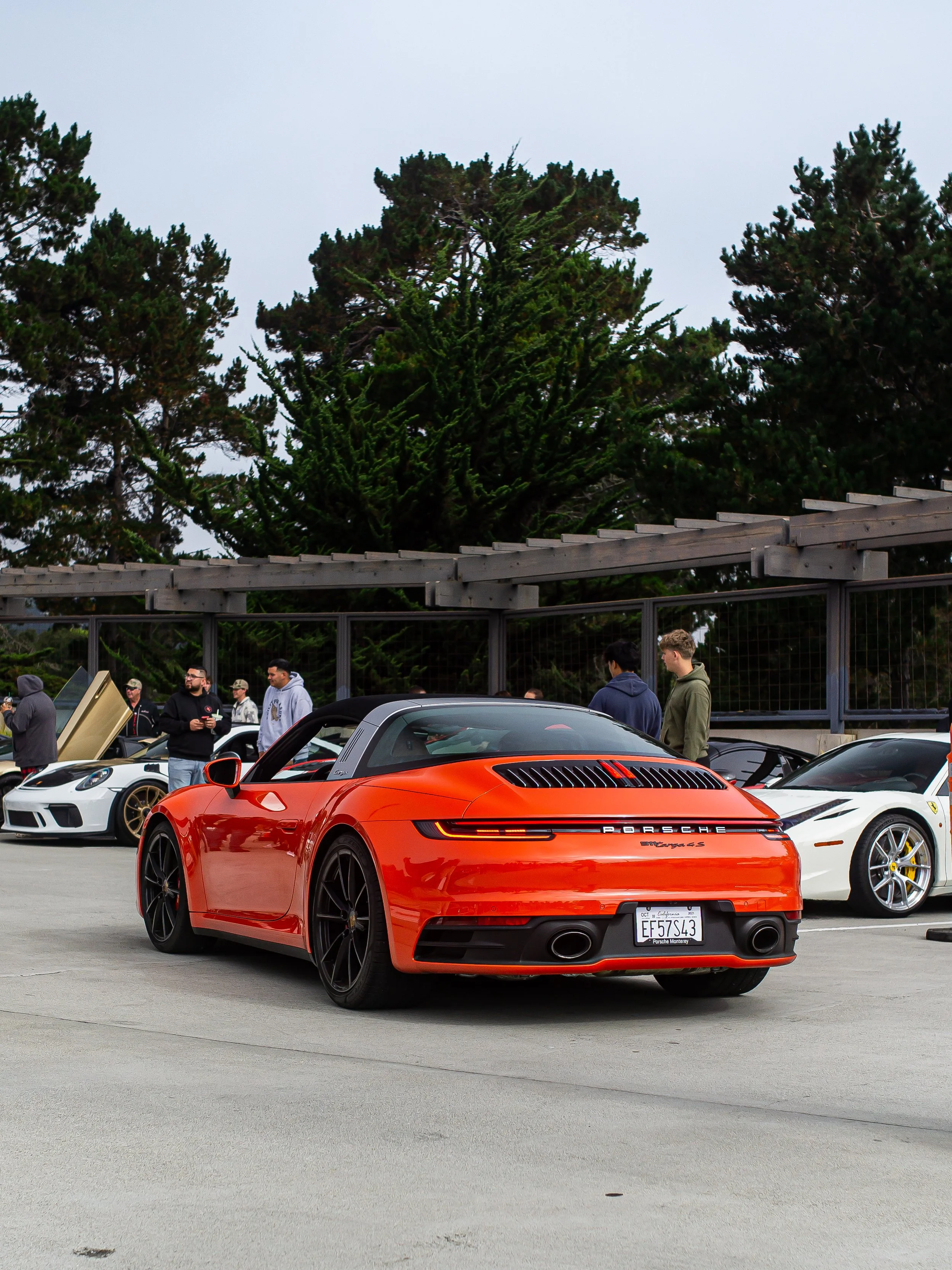 Red Porsche 911 Carrera S parked at a car meet with other sports cars and people in the background on a cloudy day.