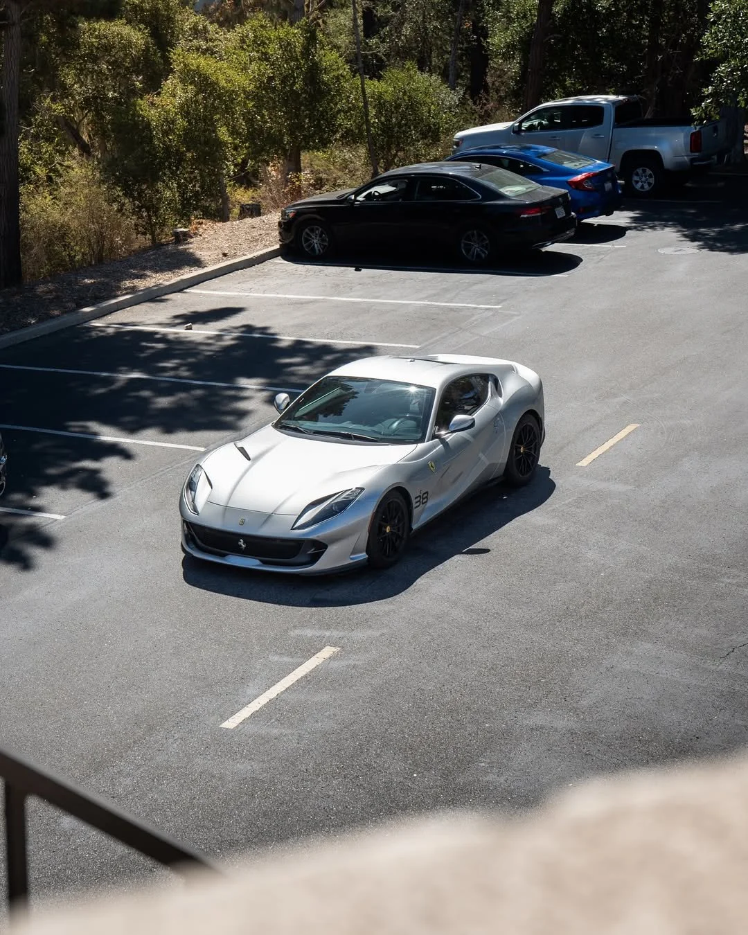 A silver Ferrari sports car parked in an almost empty parking lot with a black coupe, a blue vehicle, and a white pickup truck parked nearby, surrounded by trees and sunny weather.