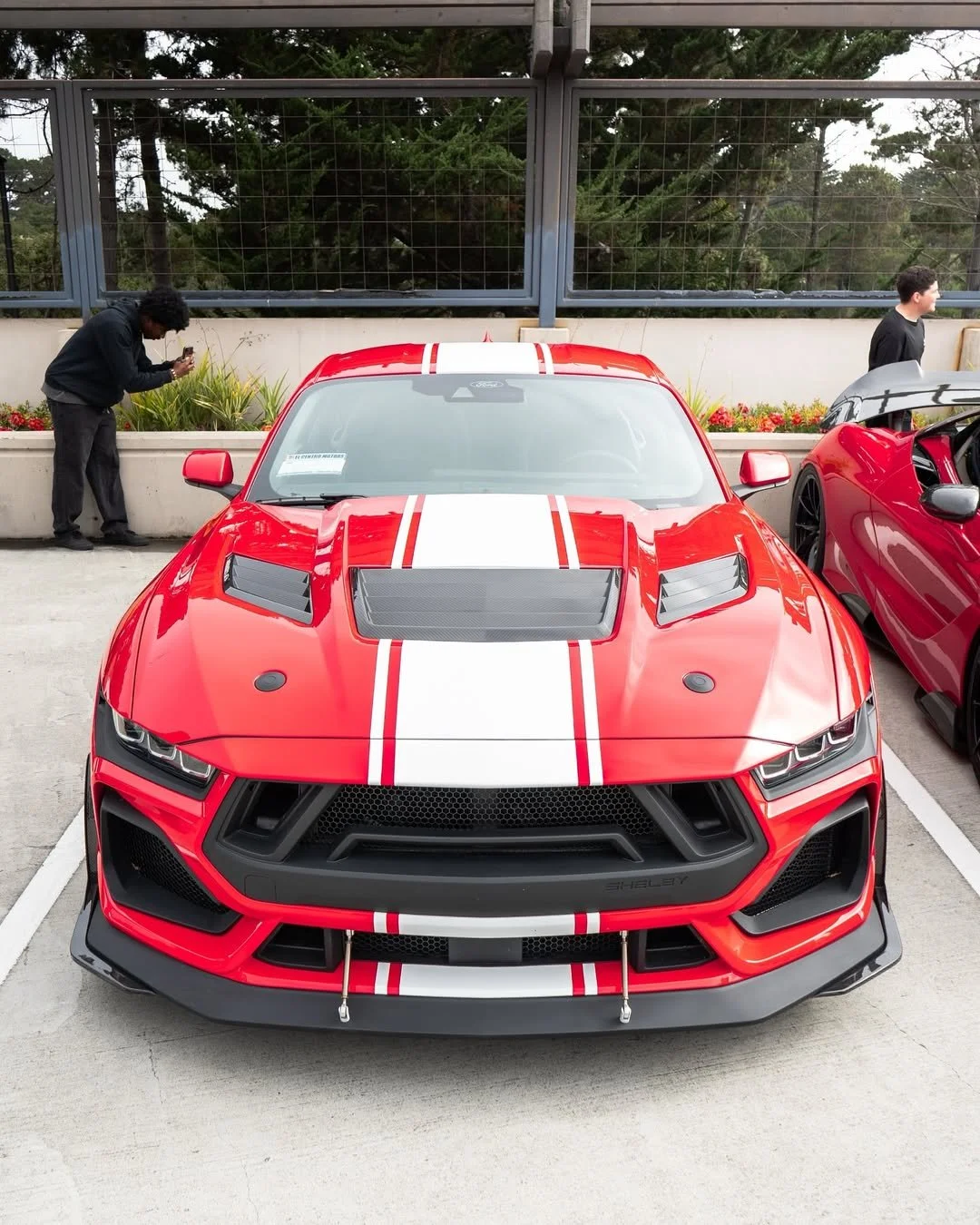 Red sports car with white racing stripes on the hood, parked in a parking lot, with two people standing nearby. One person is taking a photo, and another is looking at a car with an open door.