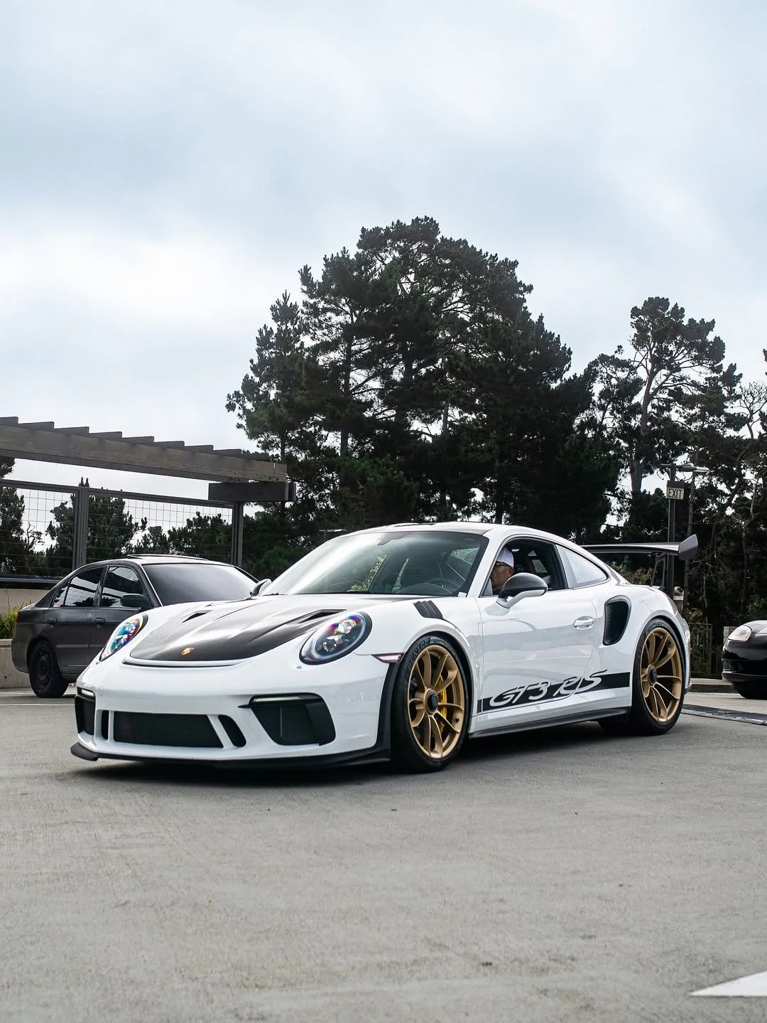 White Porsche 911 GT3 RS with gold wheels parked in a parking lot, black racing stripe on hood, side graphics, large rear wing, under cloudy sky with trees in the background.