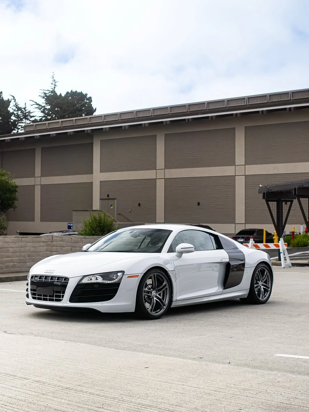 A silver Audi R8 sports car parked in a lot in front of a large building with a brown and beige wall, overcast sky.