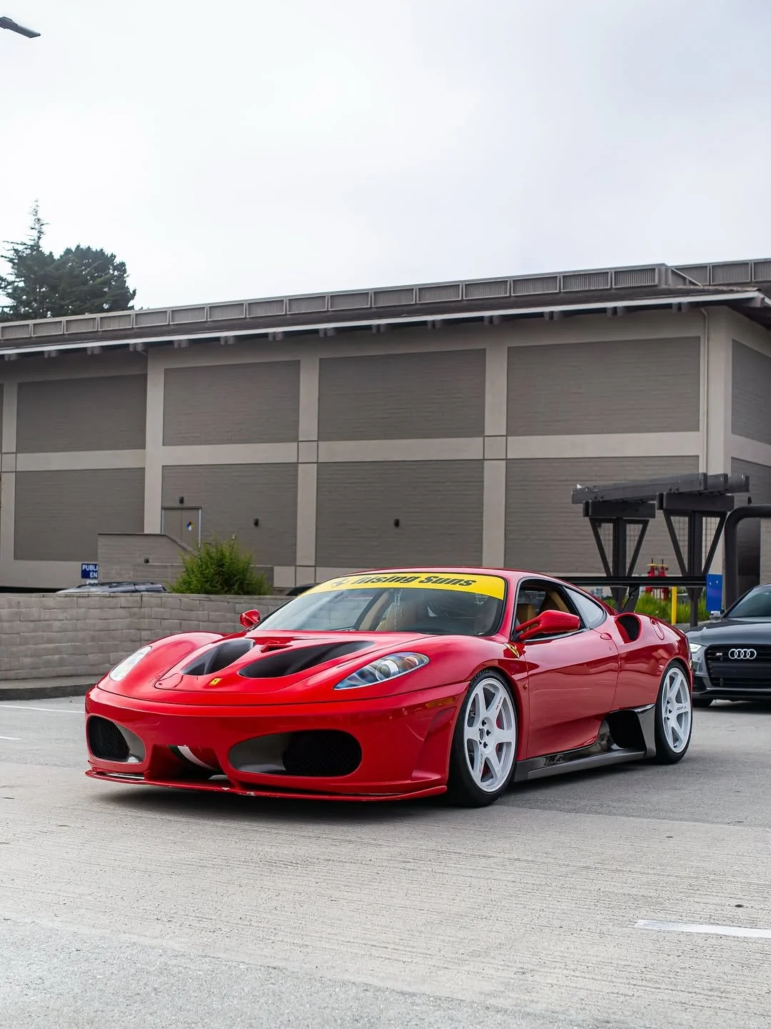 Red Ferrari sports car parked on the street with two other cars behind it and a beige building in the background.