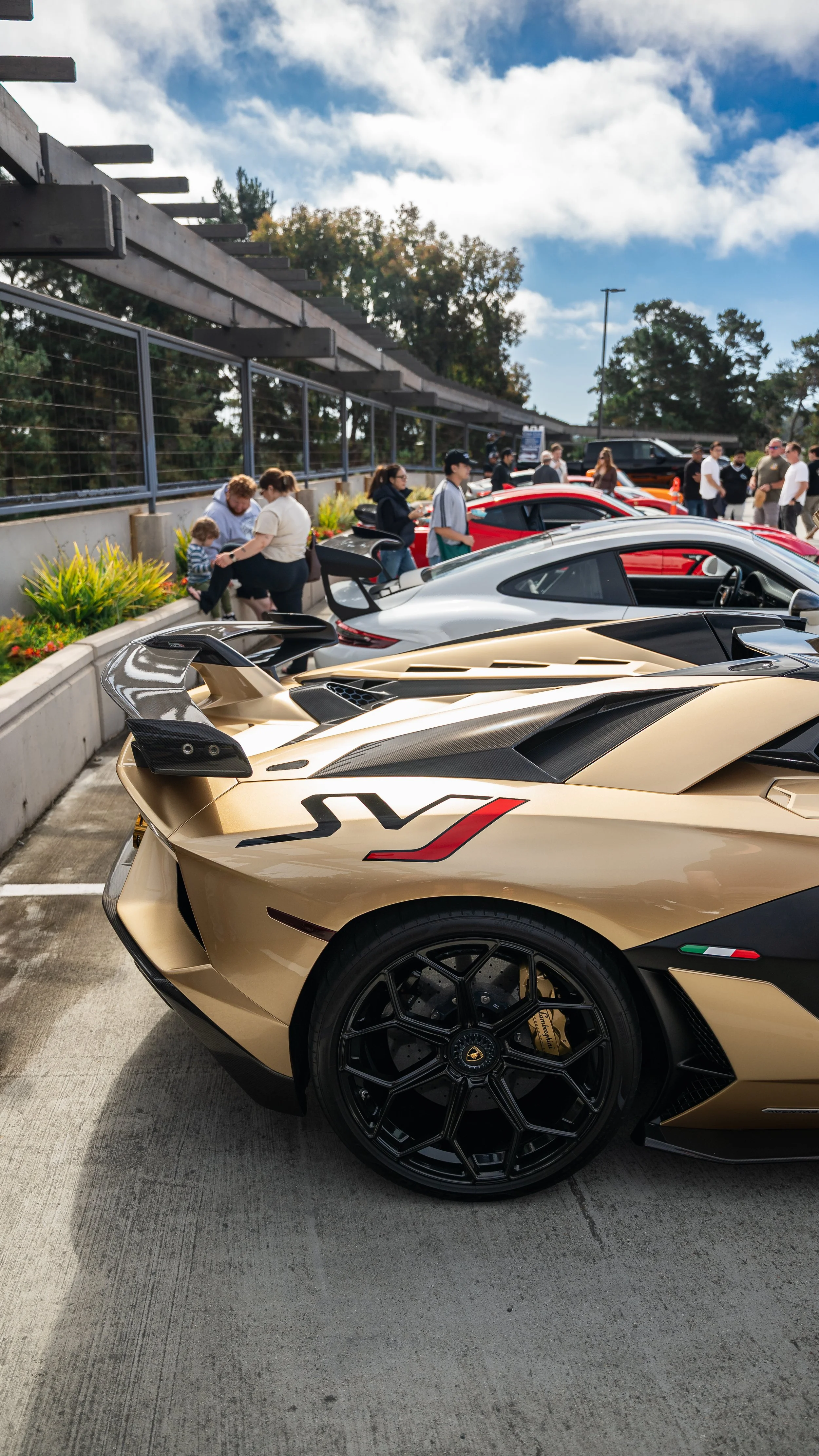 A lineup of luxury sports cars parked outdoors during a car show, with a prominent gold-colored Lamborghini and several other high-end vehicles, while people, including families and enthusiasts, view the cars under a partly cloudy sky.