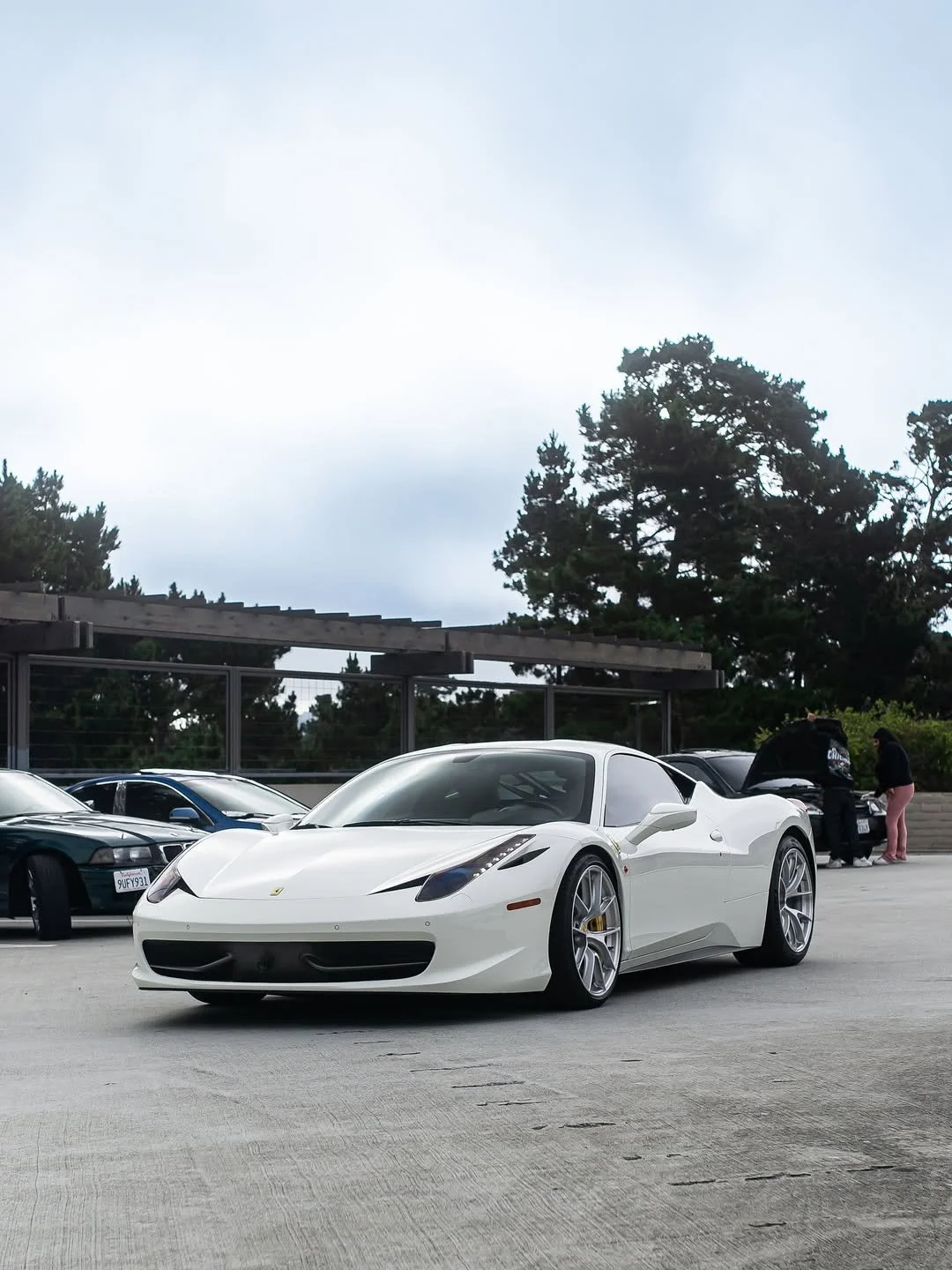 White Ferrari sports car parked in a lot with other cars, trees, and overcast sky in the background.