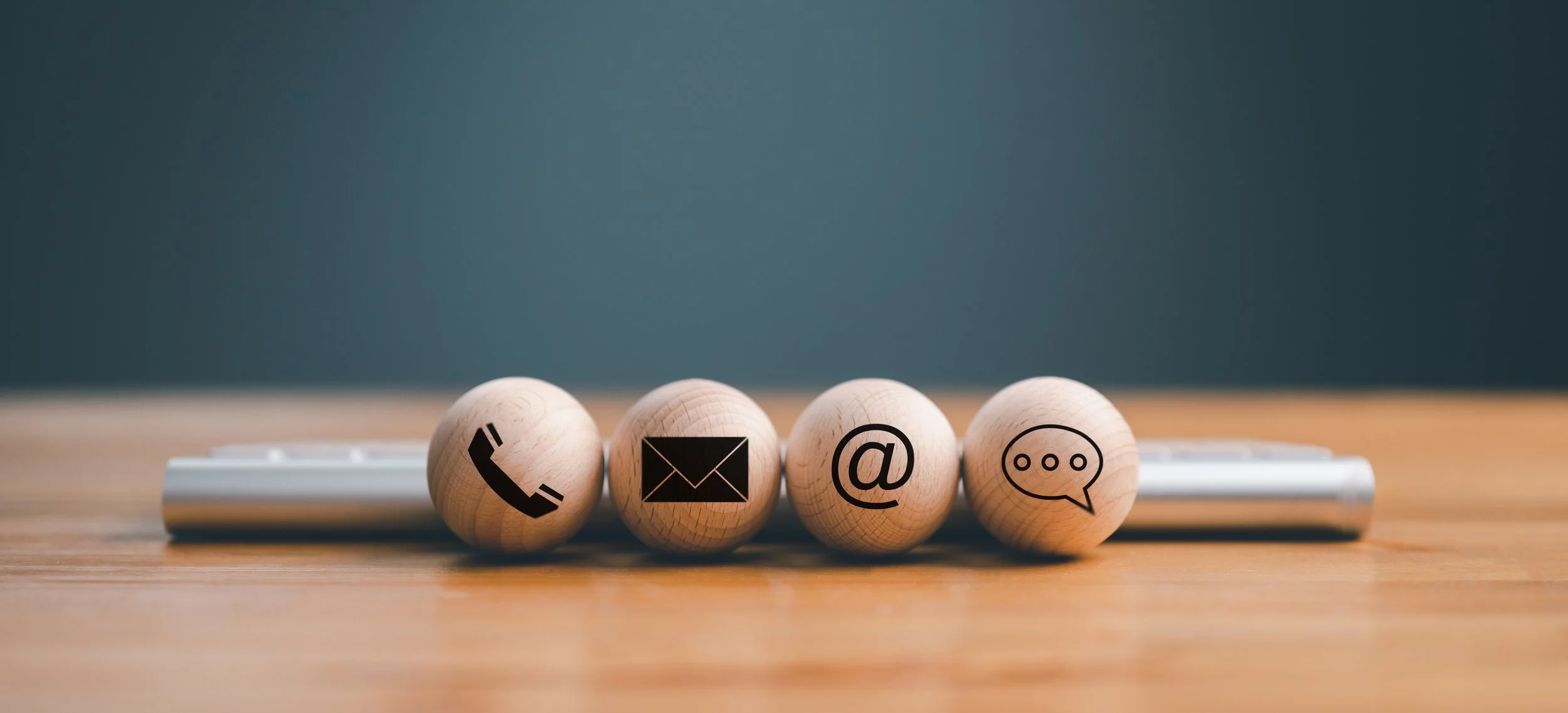 Four wooden balls with icons of a phone, email, at symbol, and chat bubble on a wooden surface.