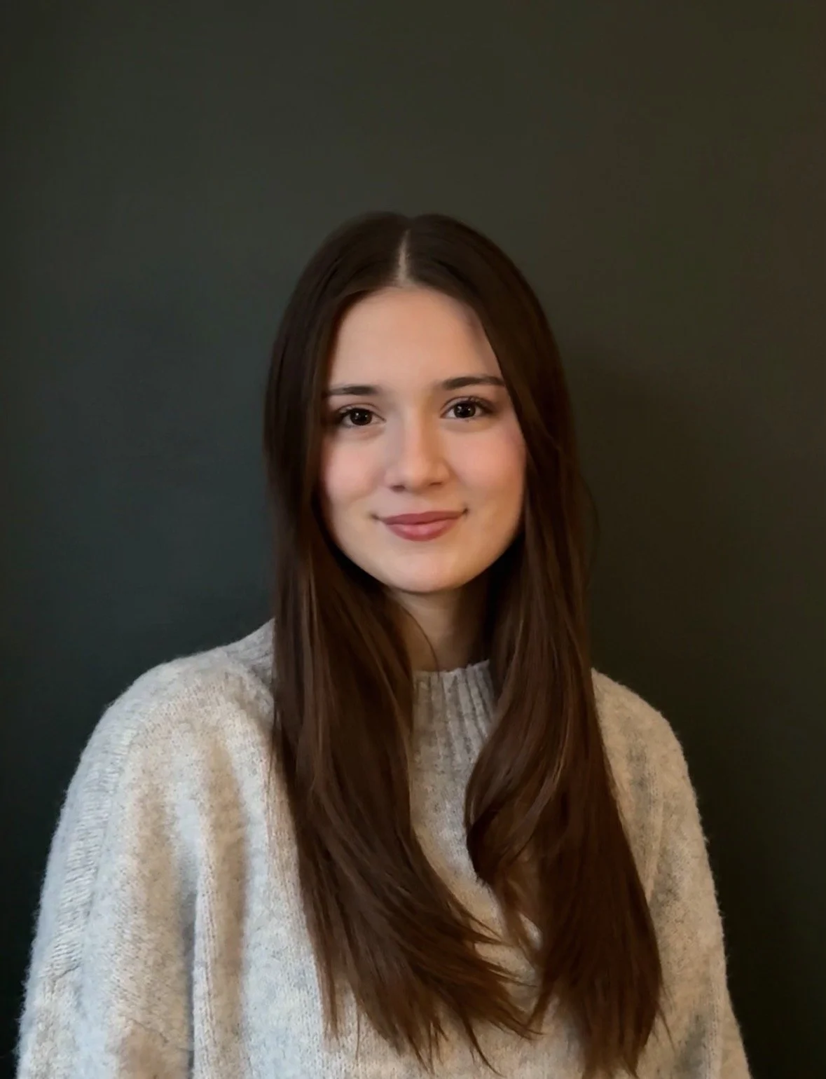 Young woman with long brown hair, smiling gently, wearing a light beige sweater, standing against a dark background.