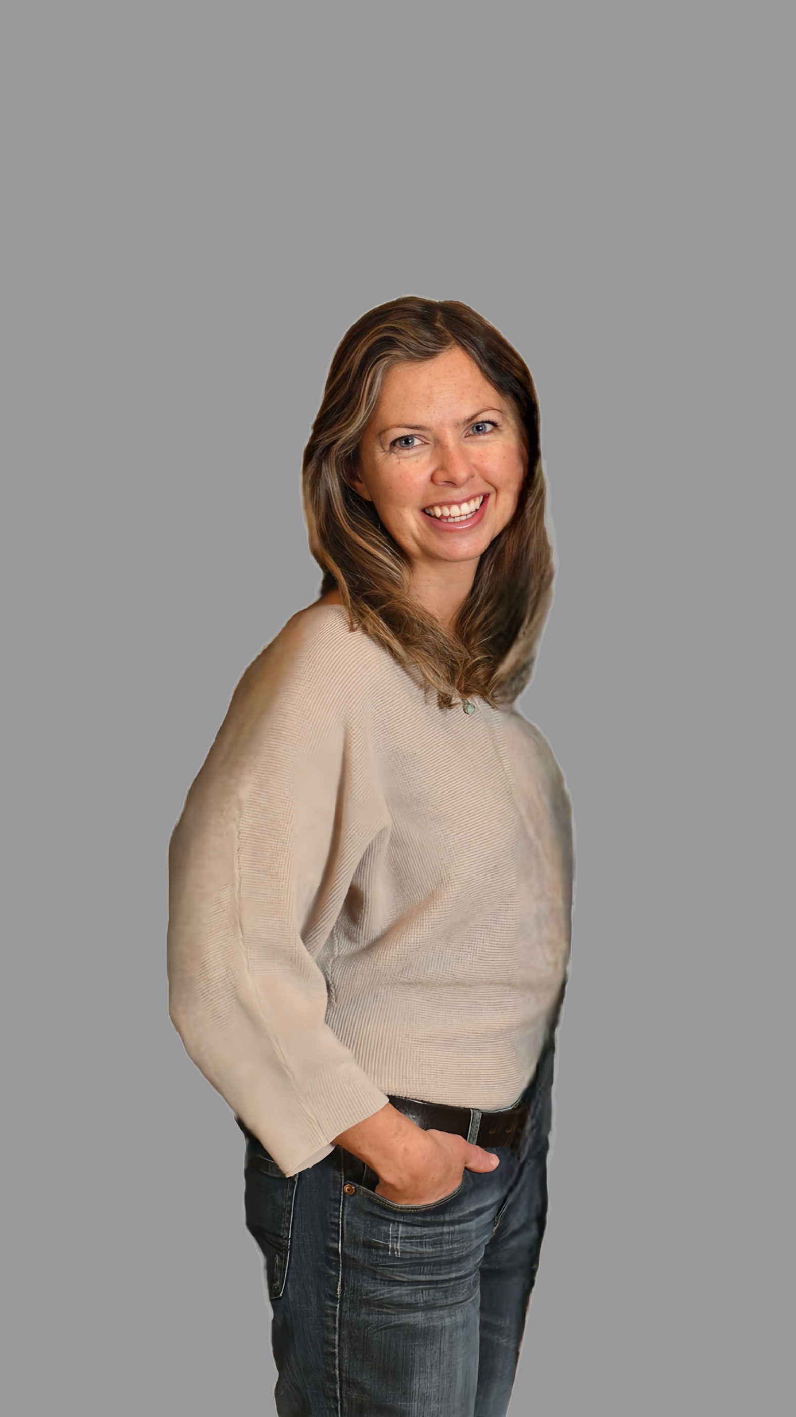 A woman with shoulder-length brown hair, smiling, wearing a beige long-sleeve shirt and dark jeans, standing against a solid gray background.