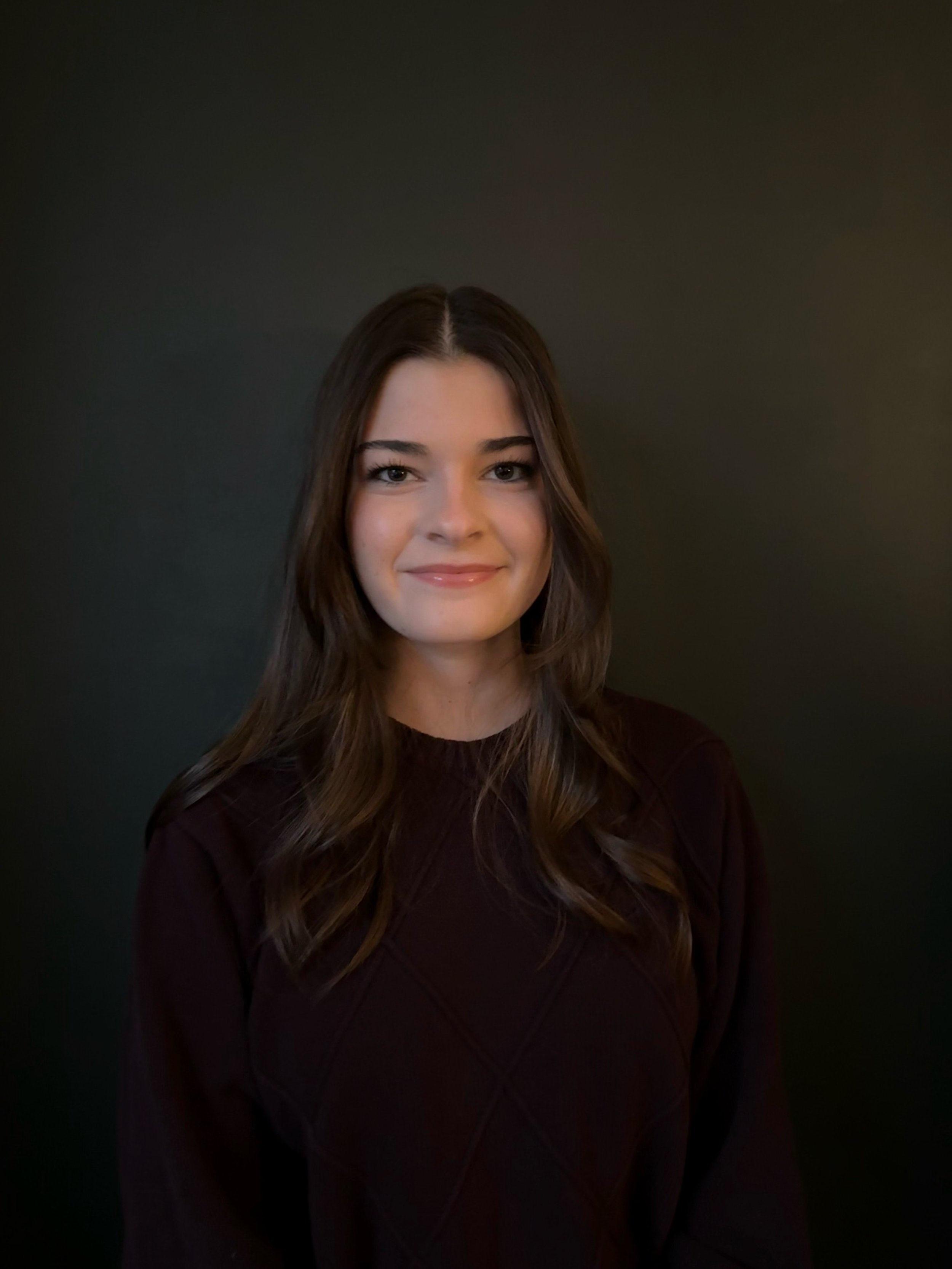 A woman with long brown hair, wearing a dark top, smiling against a dark background.