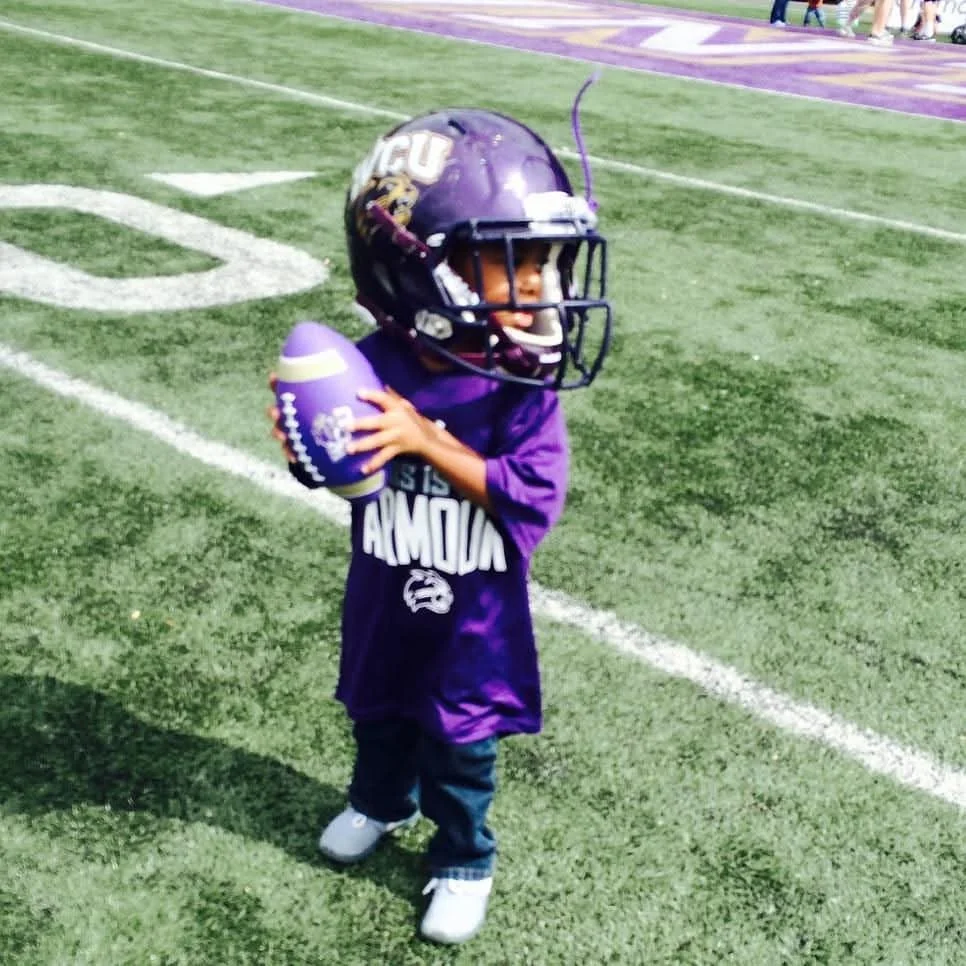 A young child standing on a football field wearing a large football helmet, holding a purple football, and dressed in a purple football jersey.