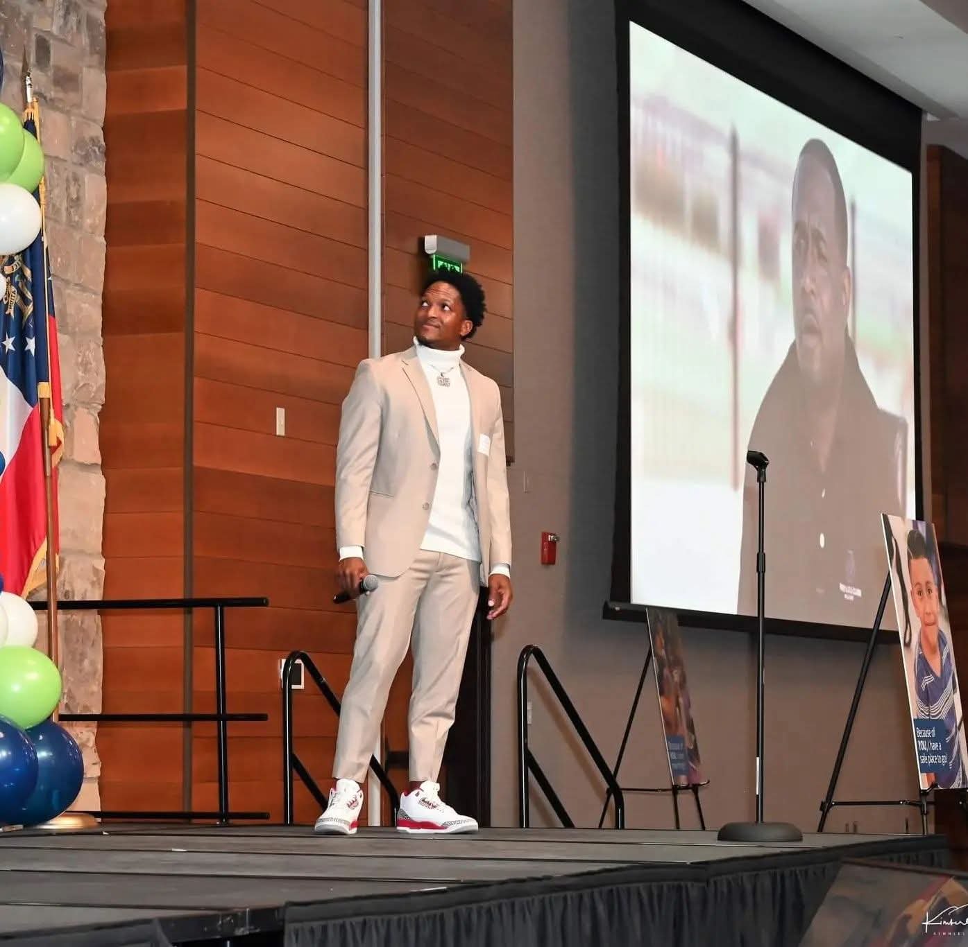 A man in a beige suit and sneakers stands on stage holding a microphone, looking up. Behind him, a large screen displays a video of a woman speaking. There are posters and balloons on the stage, and a flag to the left.