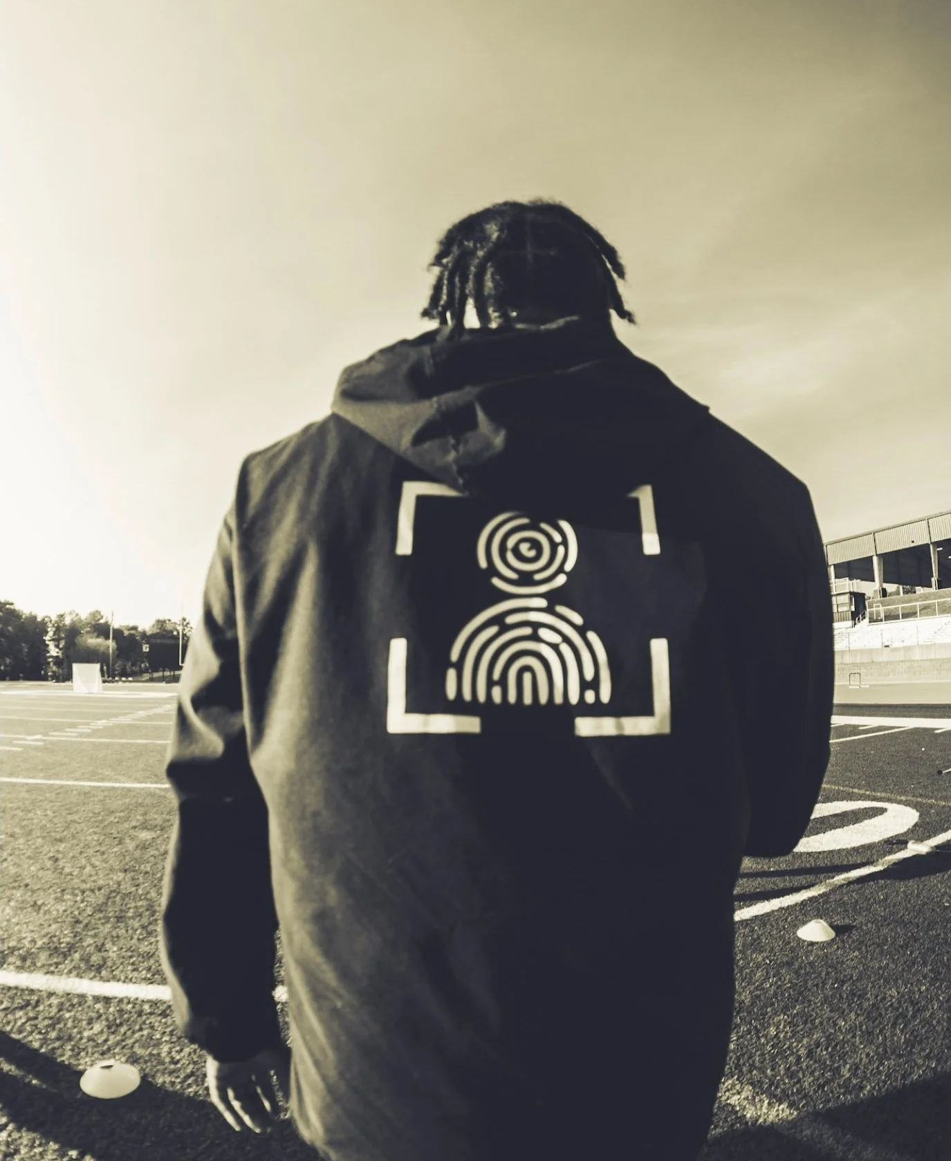 A person with dreadlocks wearing a jacket with a fingerprint design on the back, standing on an outdoor sports track.