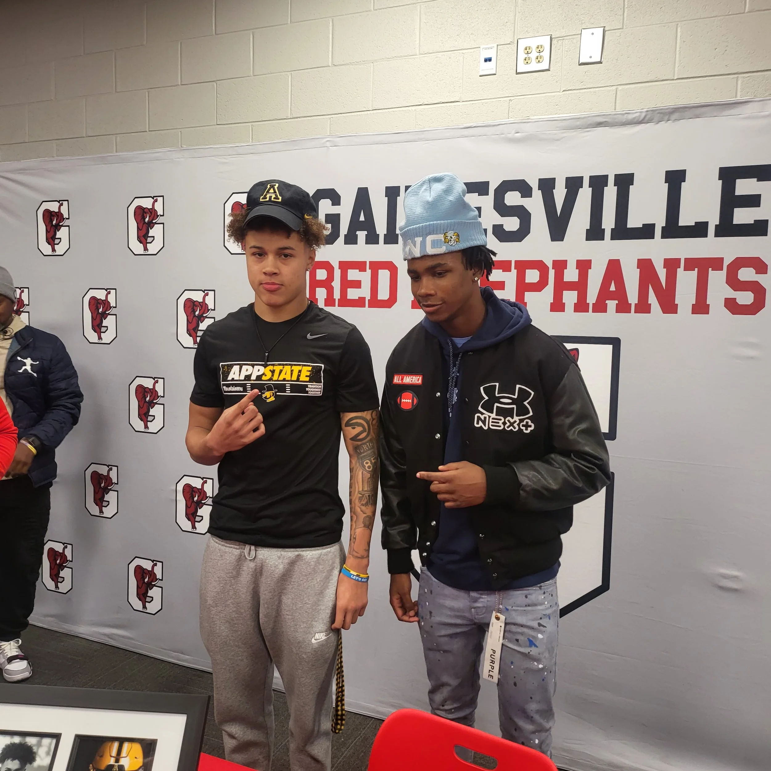 Two young men standing in front of a Gainesville Red Elephants banner. The young man on the left is wearing a black Nike T-shirt, gray sweatpants, a black cap with yellow A, and is making a hand gesture. The young man on the right is wearing a black 