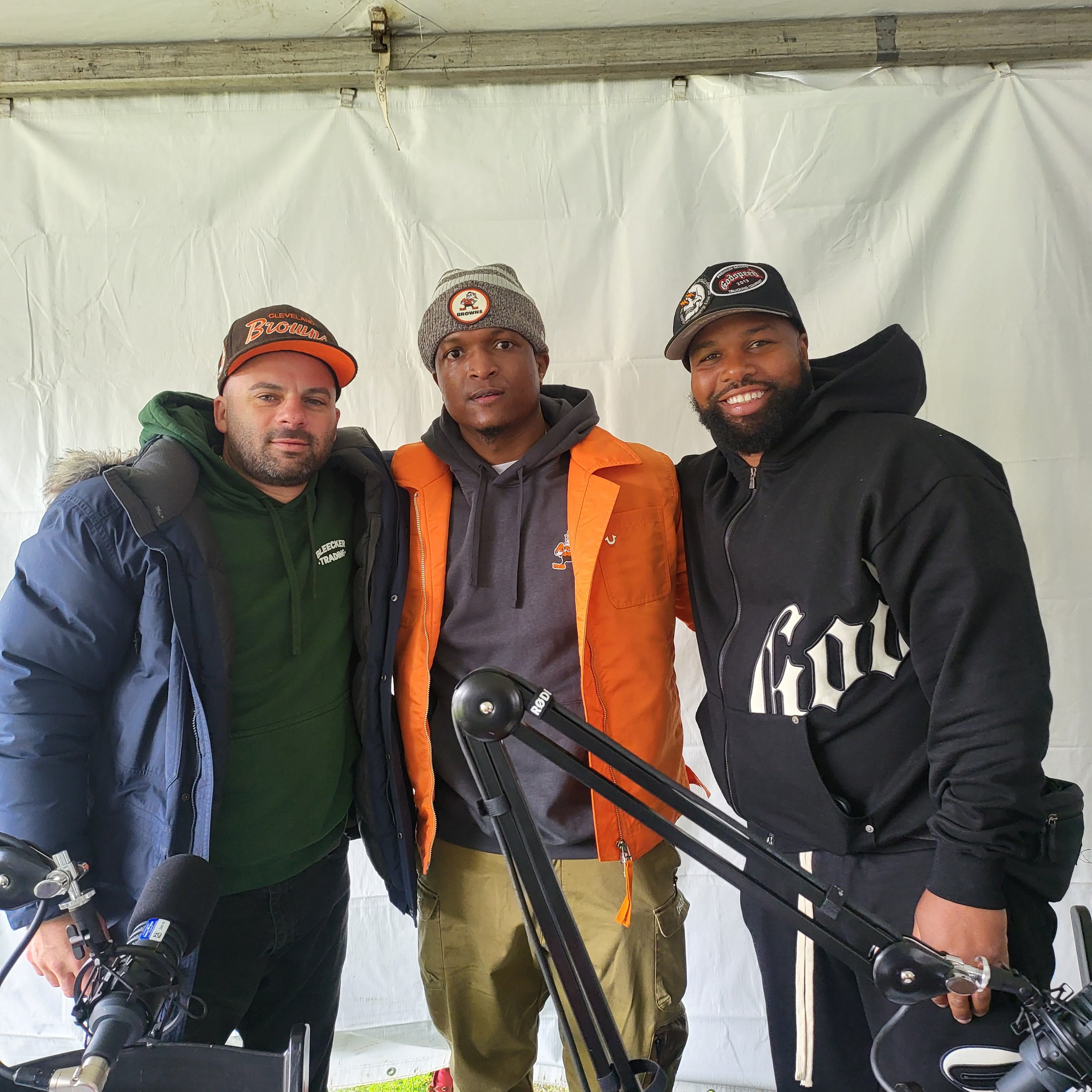 Three men posing for a photo behind a bicycle, standing under a white canopy. The man on the left is wearing a gray cap with orange accents and a green jacket, the man in the middle is wearing a gray beanie with a Cleveland Browns logo and an orange 