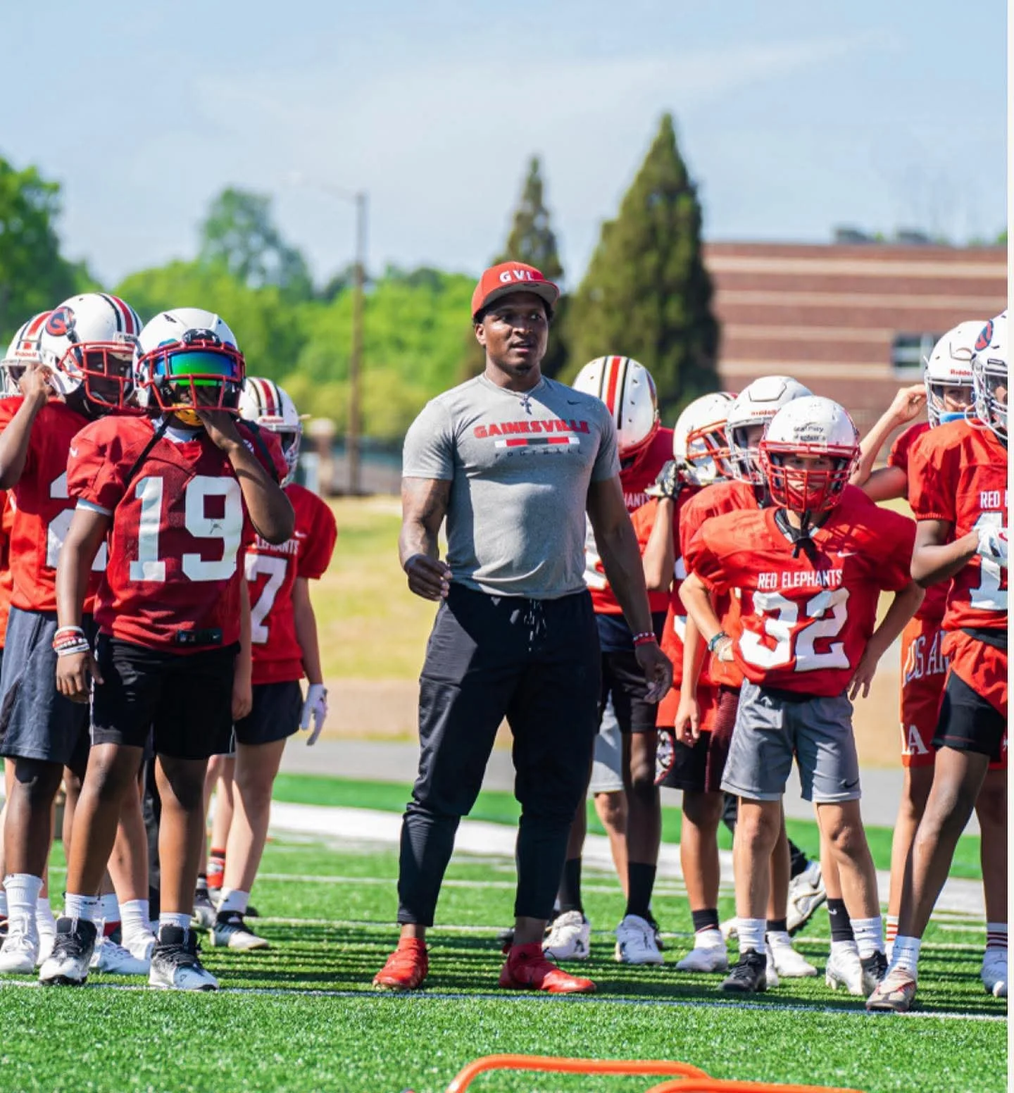 A football coach stands on a field with a group of young players in red practice jerseys and helmets during a practice session.