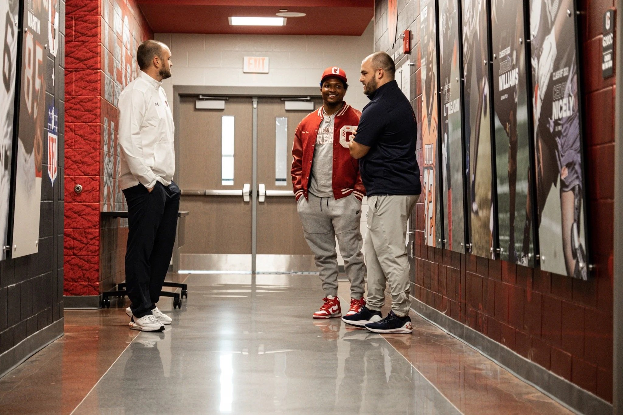 Three men standing and talking in a hallway near a double door with posters on the wall.