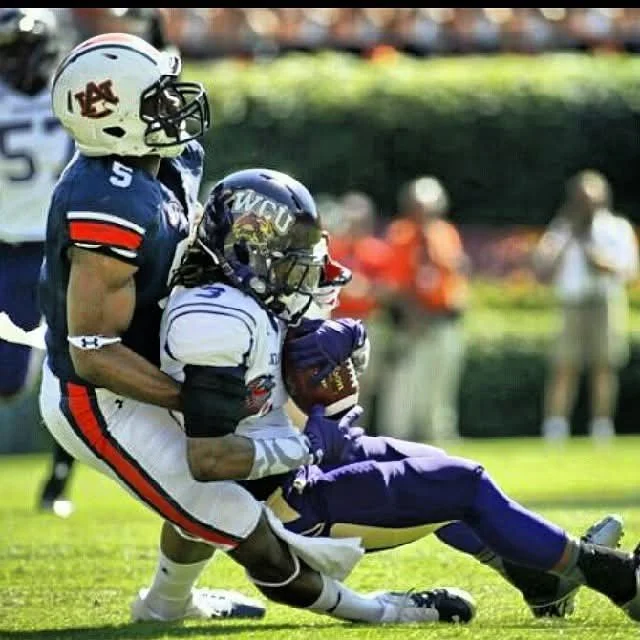 Two football players in helmets and uniforms compete for the ball during a game on a grass field, with spectators in the background.