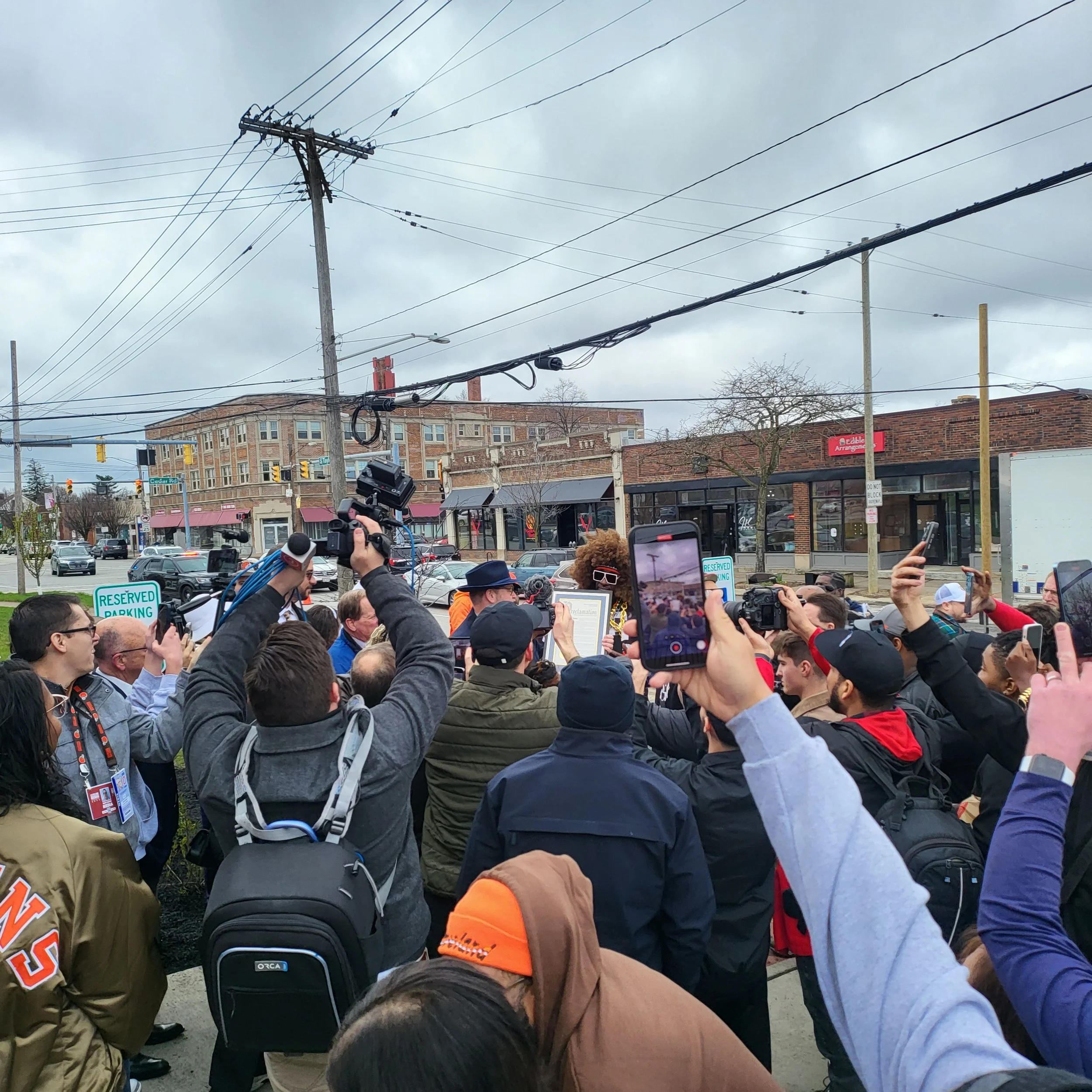 Crowd of people gathered outdoors on a city street. Many are taking pictures or recording with smartphones and cameras. The sky is overcast, and there are buildings, telephone poles, and traffic lights in the background.