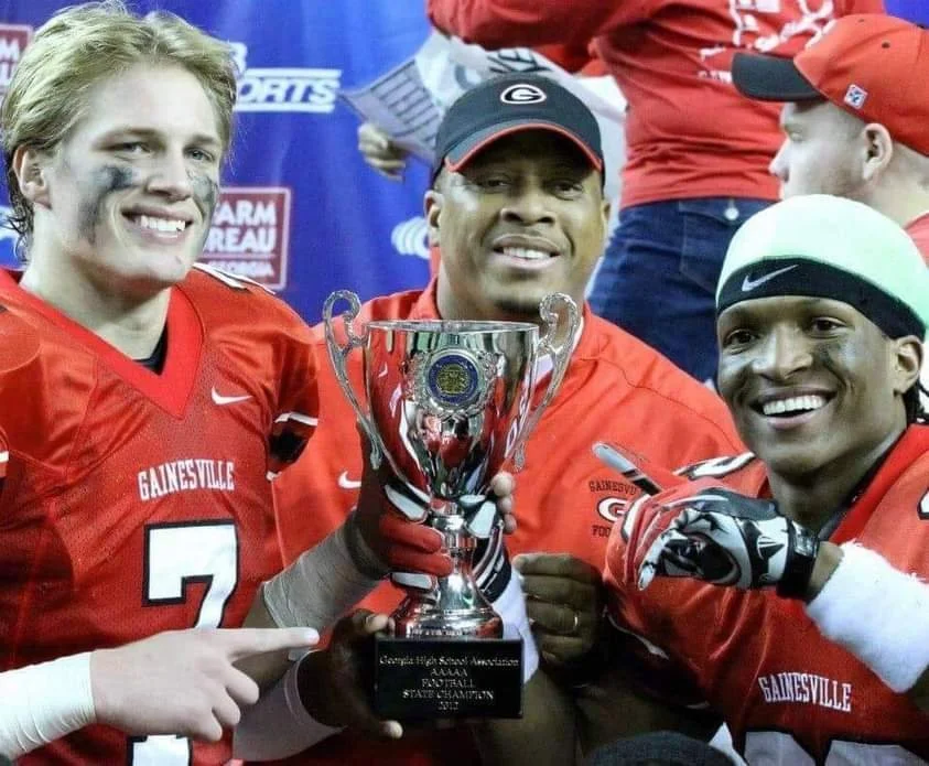 Three football players in red uniforms celebrating with a silver trophy after a game, smiling.
