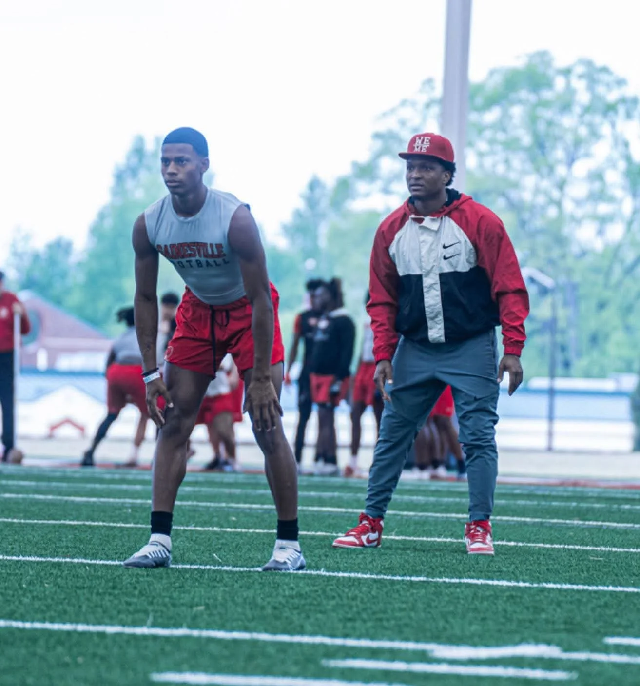 A young male athlete in a gray sleeveless shirt and red shorts standing on a running track, with a coach or trainer in a red and gray jacket and red cap standing nearby. The background includes other athletes and a sports field.