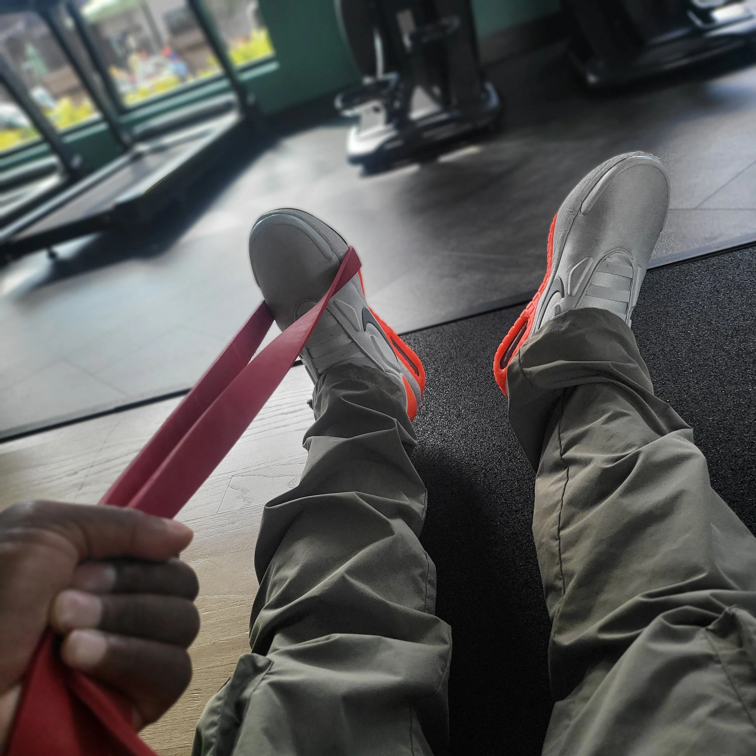 Person wearing gray and red sneakers and khaki pants sitting indoors, holding a red resistance band, with gym equipment and windows in the background.