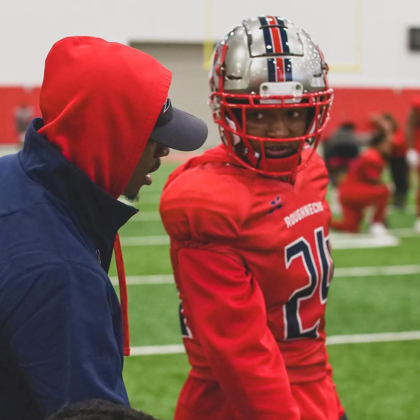 A football coach and a player engaged in conversation on a field, with the player wearing a red uniform, helmet, and shoulder pads, and the coach wearing a navy blue jacket with a red hood and a gray cap.