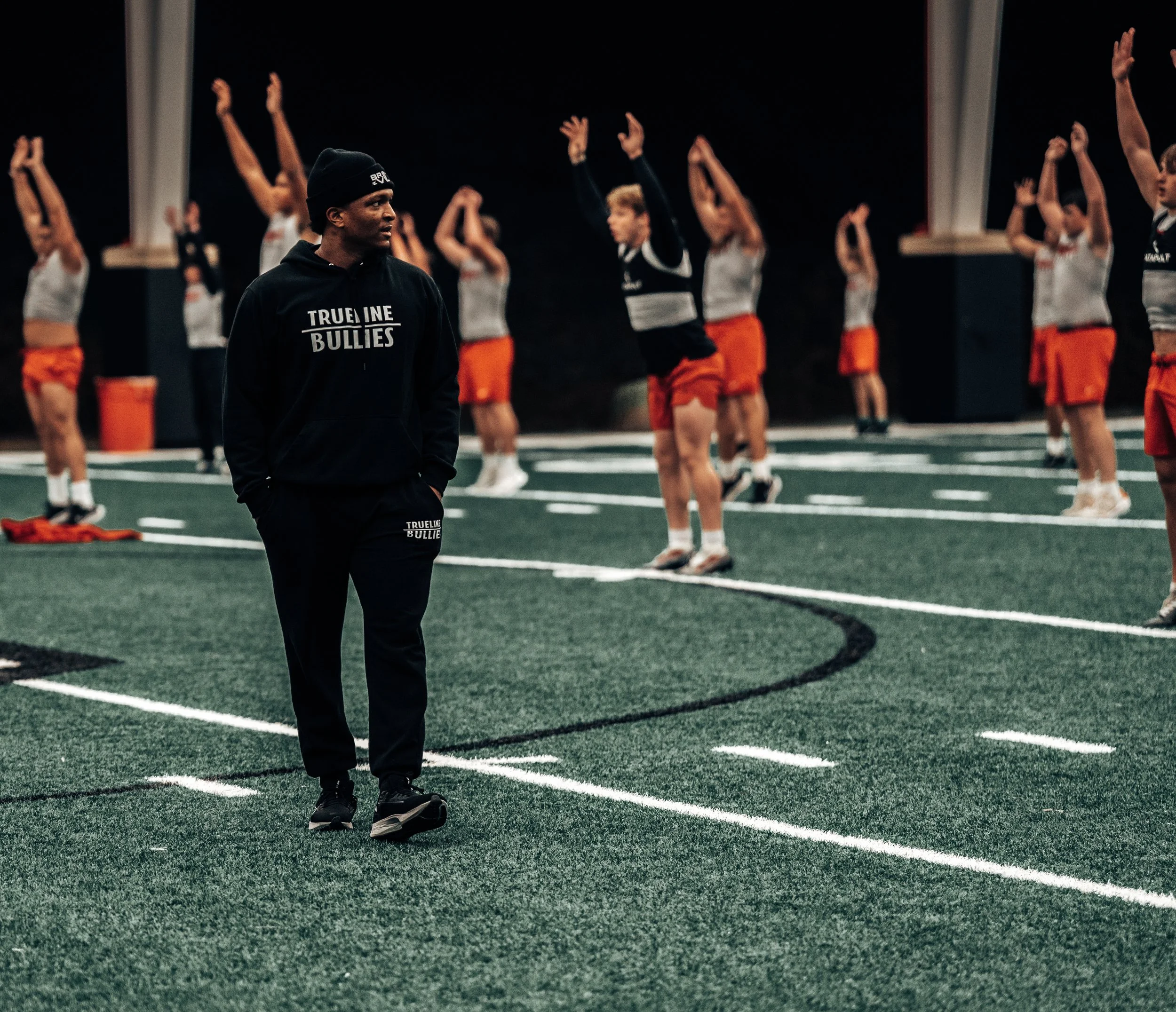 A coach standing on a gymnasium turf watching a group of young female athletes stretching and warming up in sports uniforms.