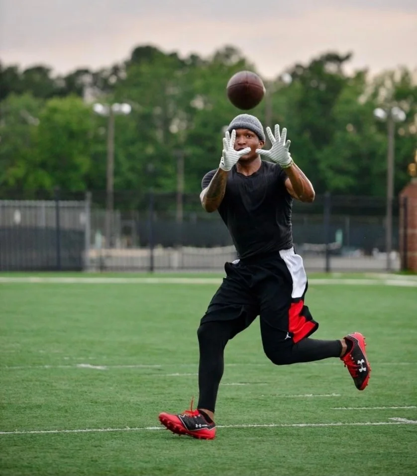 A football player on a field preparing to catch a football, with an athletic stance, wearing gloves, a beanie, and sportswear under an overcast sky.