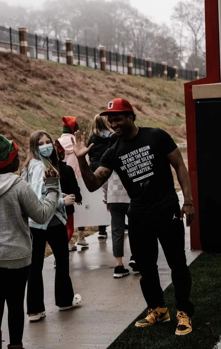 Man high-fiving children at an outdoor event; man is wearing a black T-shirt with an inspirational quote, a red cap, and yellow and black sneakers; children are wearing face masks and winter clothing; background shows a grassy hill with a fence, and 