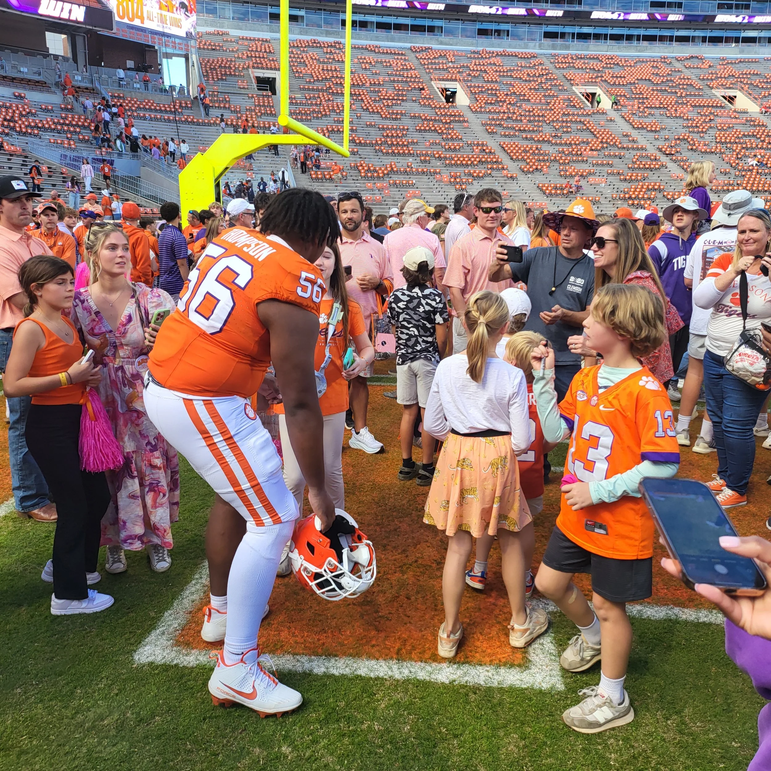 A football player wearing an orange and white uniform with the number 56 on the jersey stands on the field, surrounded by fans including children and adults, some taking photos, with the stadium seating in the background.