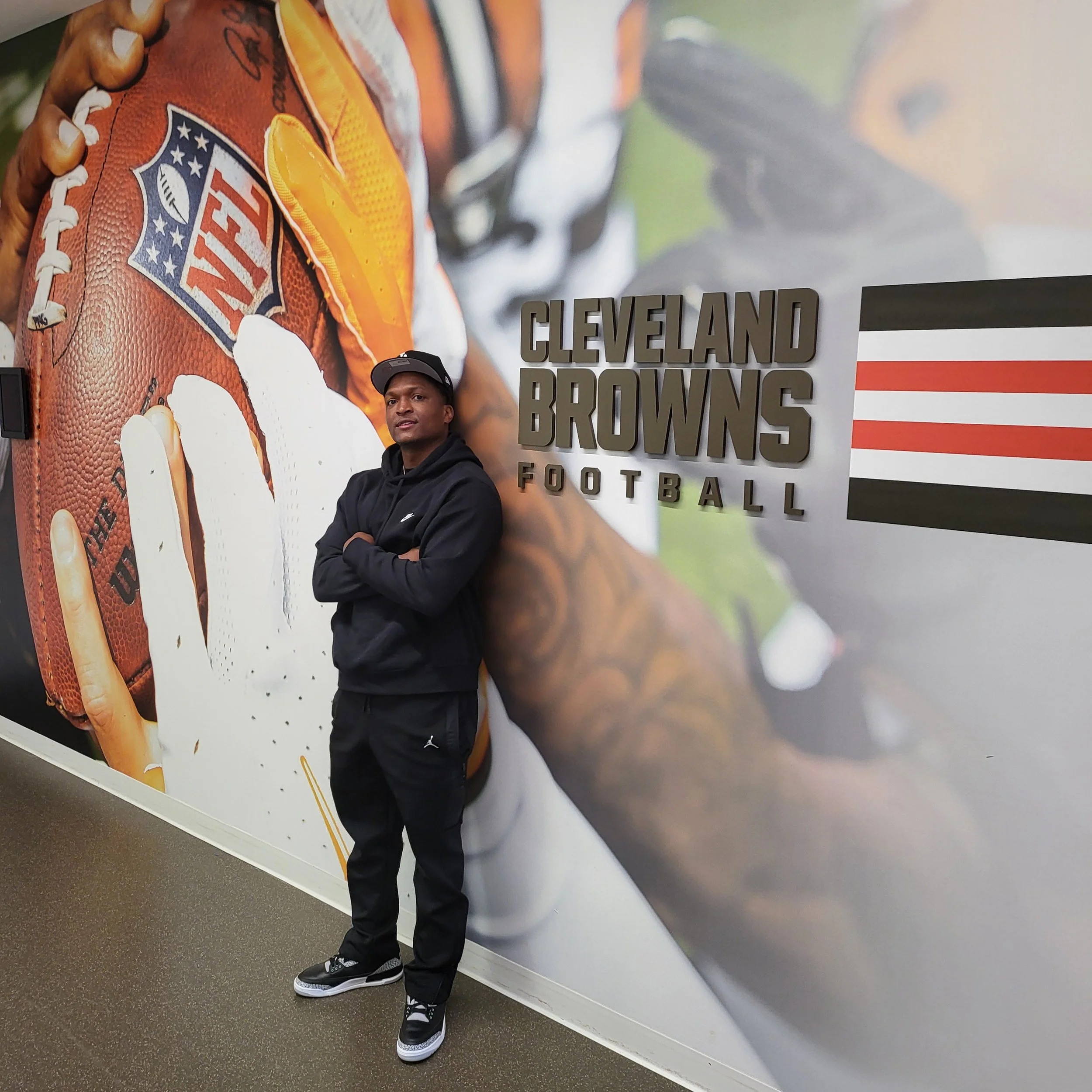A man with dark skin wearing a black hoodie, black pants, black and white Nike sneakers, and a gray Nike cap stands with arms crossed in front of a large Cleveland Browns football team poster at a sports event.