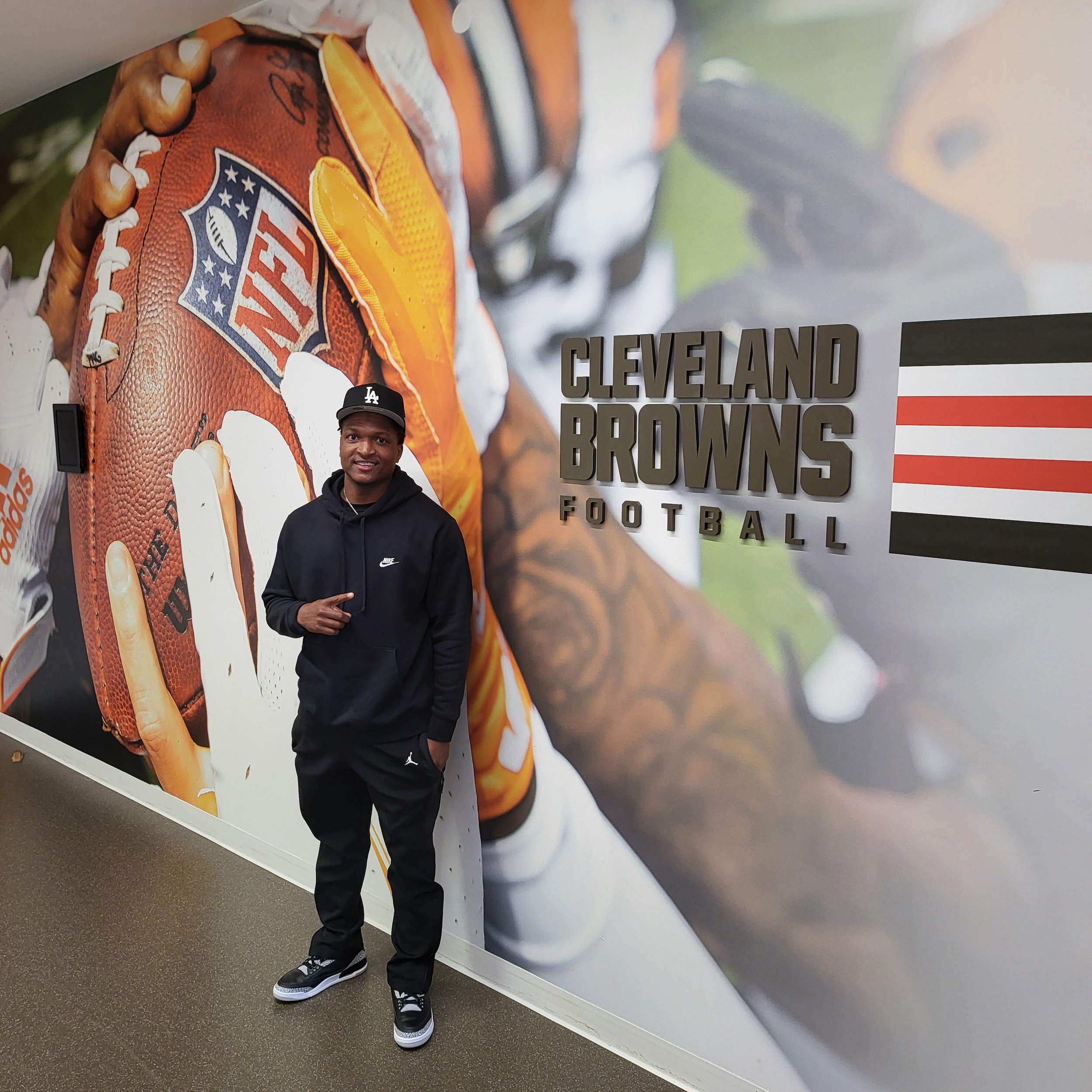 Young man standing in front of a large advertisement for Cleveland Browns football team, featuring a close-up of a football and a player in a Browns uniform, with text that reads 'Cleveland Browns Football'.