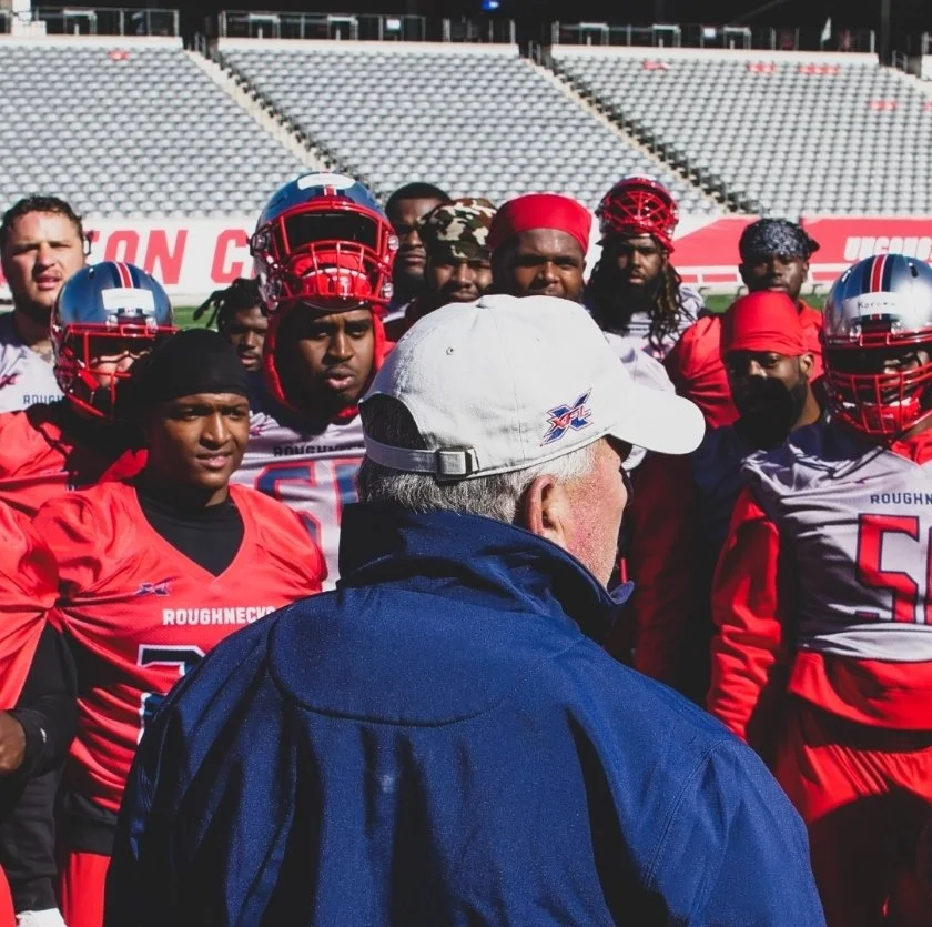 Football coach speaking to team on field with players wearing red and gray uniforms, in a stadium.