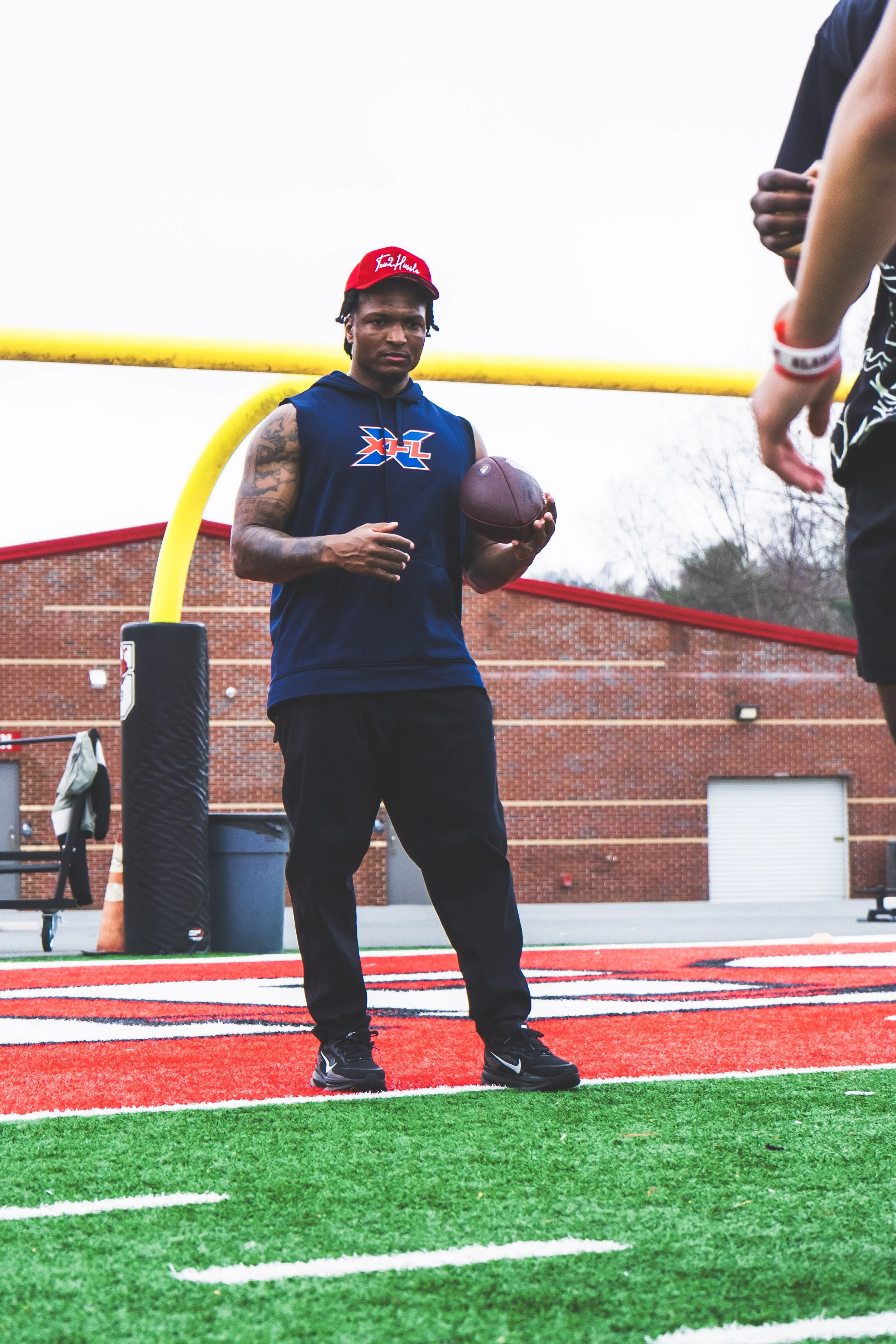 A man standing on a football field holding a football, wearing a sleeveless hoodie and a red cap, with others partially visible nearby.