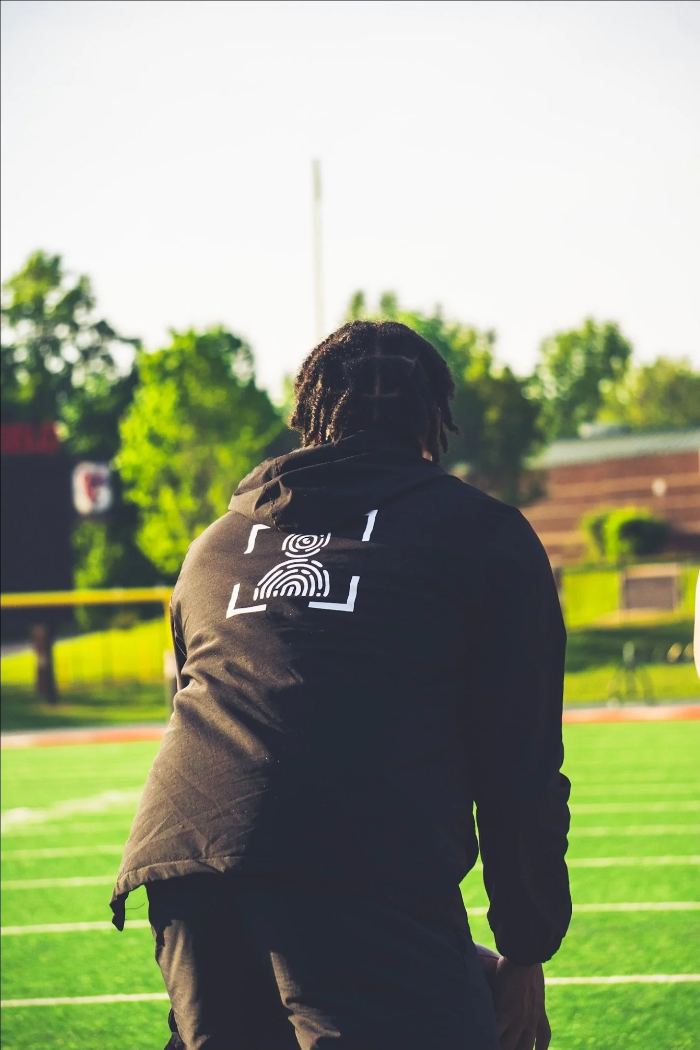 Person with dreadlocks wearing a black hoodie with white text and design, standing with their back to the camera on a grassy sports field during daytime, with trees and structures in the background.