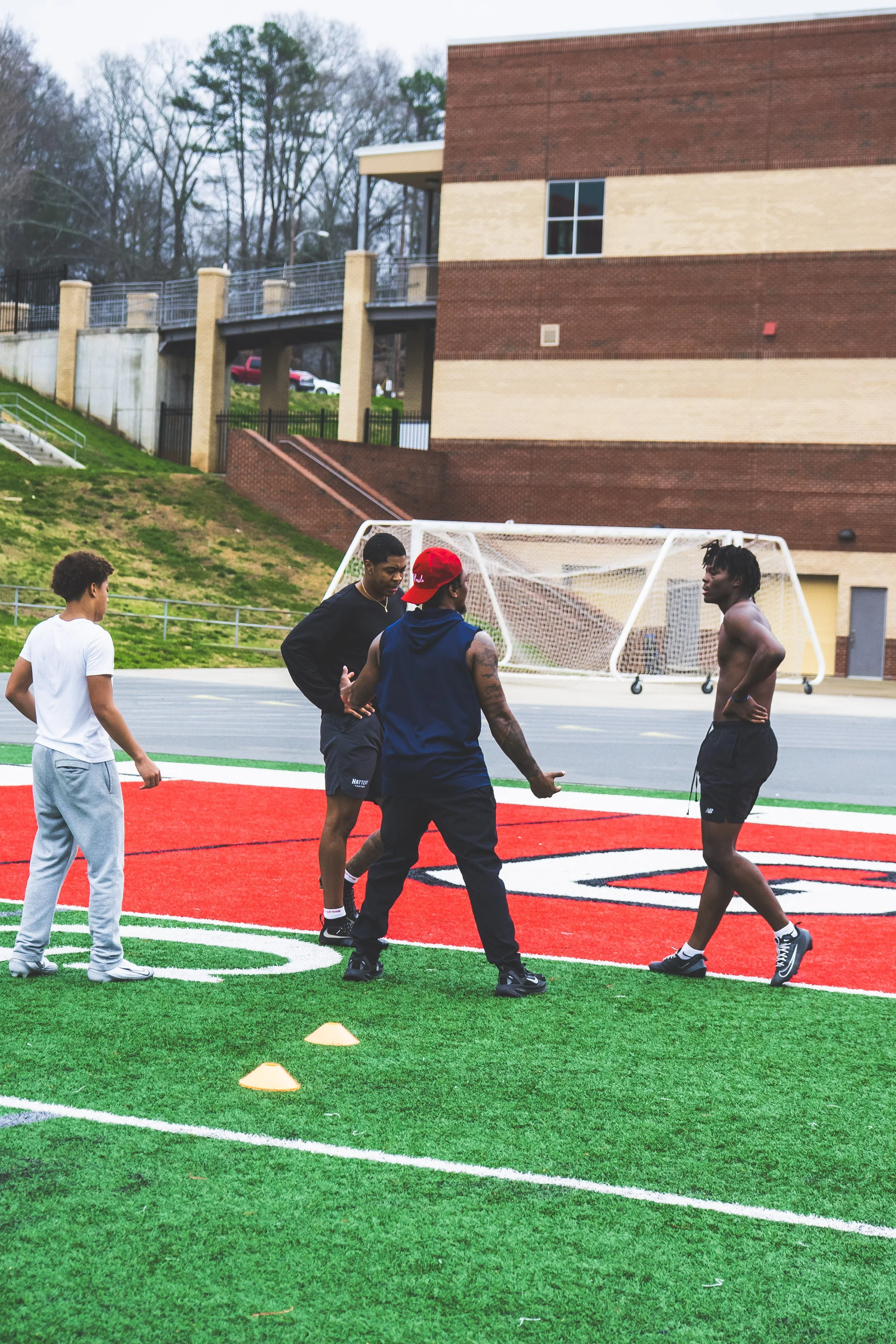 Four young men and a boy on a turf football field near a school football goal, with a large school building and trees in the background, engaging in a discussion or training.