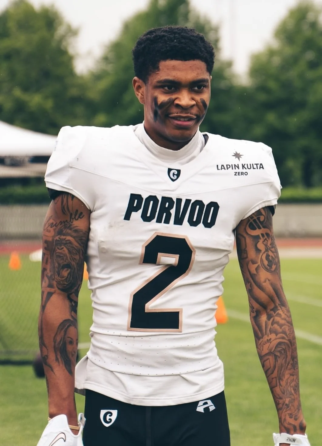A young male football player wearing a white jersey with black and gold accents, number 2, with tattoos on both arms, standing on a football field with trees in the background.