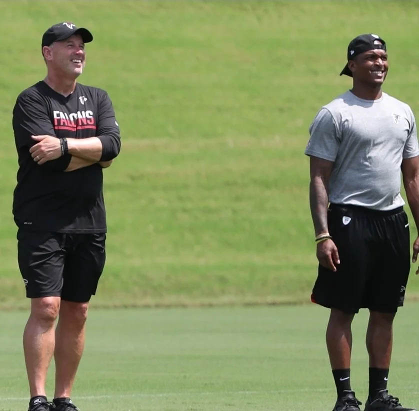 Two men standing on a grassy field, smiling and laughing. They are wearing casual athletic clothes and hats, with a relaxed outdoor setting.