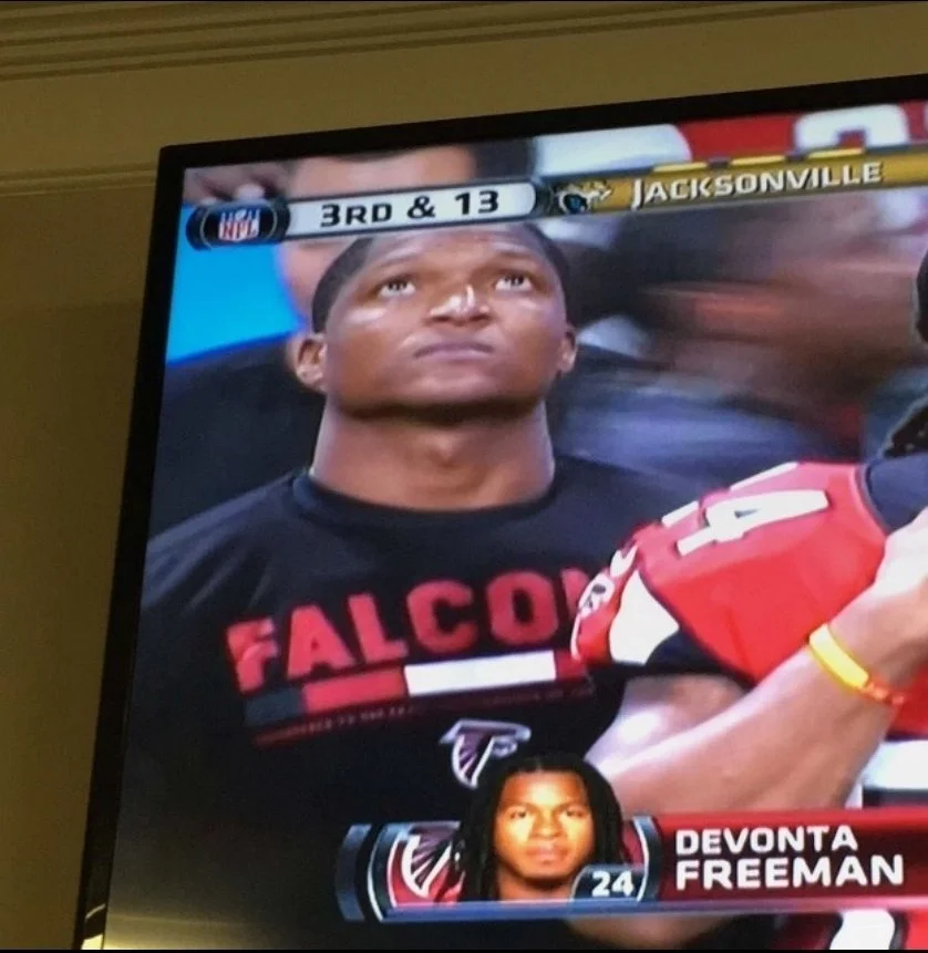 Football player from the Atlanta Falcons, Devonta Freeman, standing with a focused expression during a game, wearing black warm-up gear with red lettering, on a scoreboard screen showing game details between Jacksonville and Atlanta.