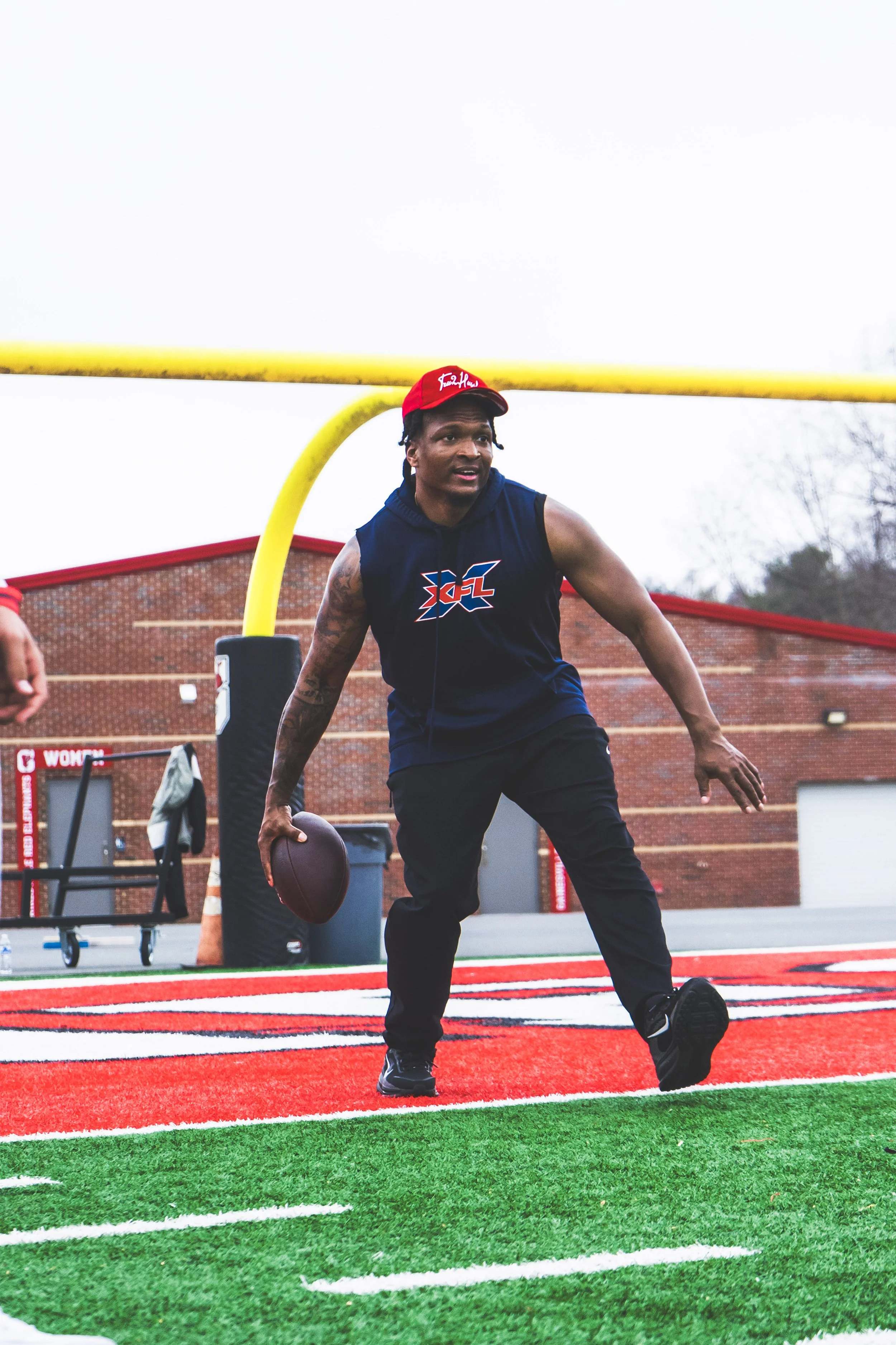 A man holding a football in a football field, standing near a yellow goal post, wearing a navy hoodie with a logo, black pants, and a red cap.