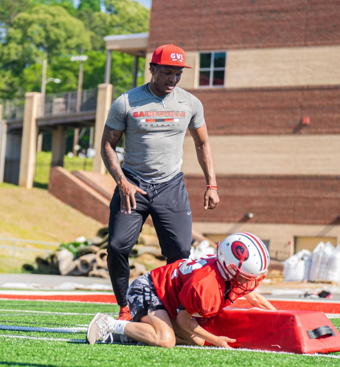 A football player in a red uniform kneeling on the field next to a red blocking pad, with a young man standing nearby. The young man is wearing a gray Gainesville football t-shirt, a red cap, and black athletic pants, observing the practice.